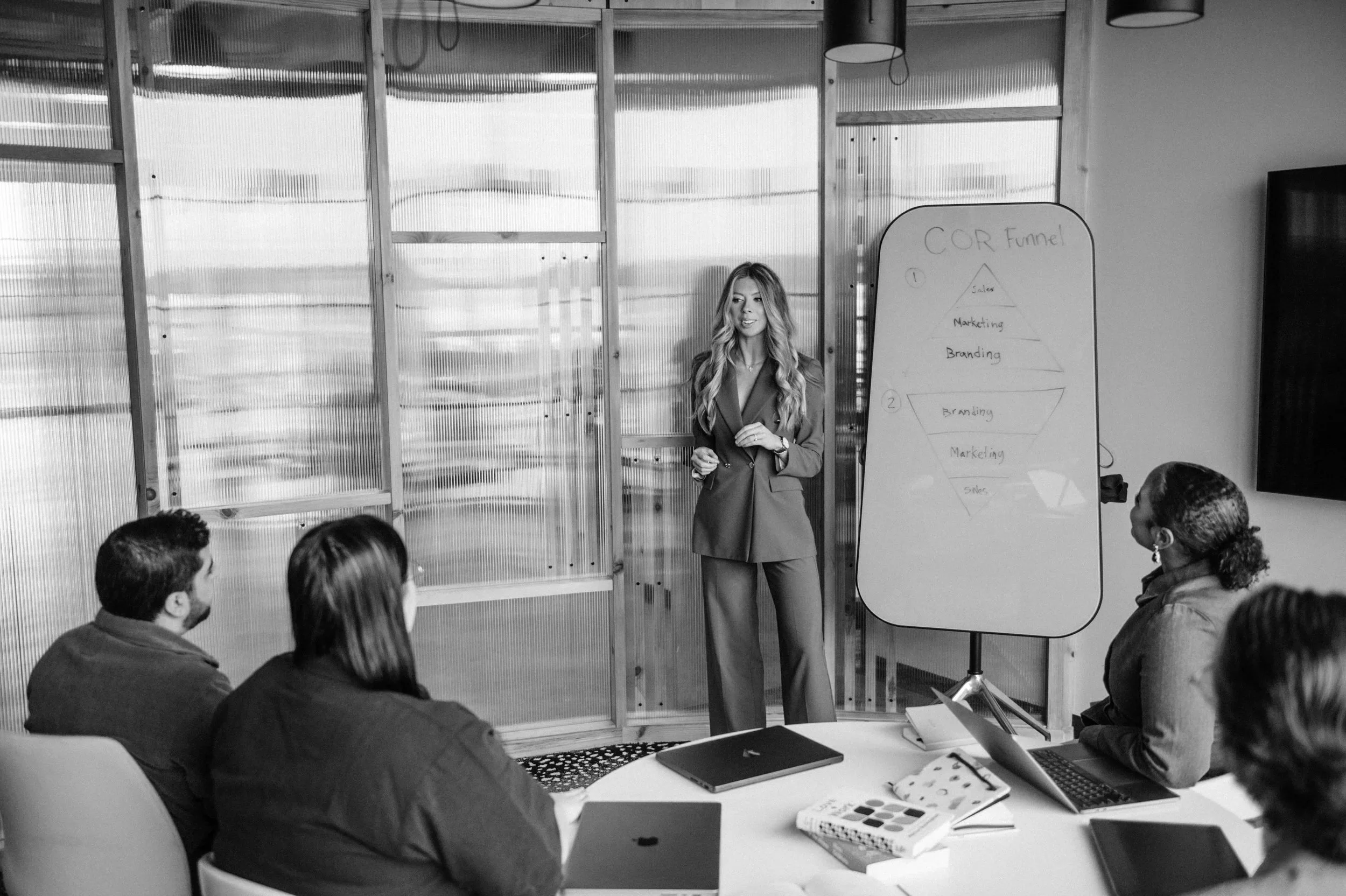 A woman standing in front of a group of people, giving a presentation in a meeting room. There is a whiteboard with a diagram and some writing behind her. The attendees are seated around a table with laptops and notebooks.