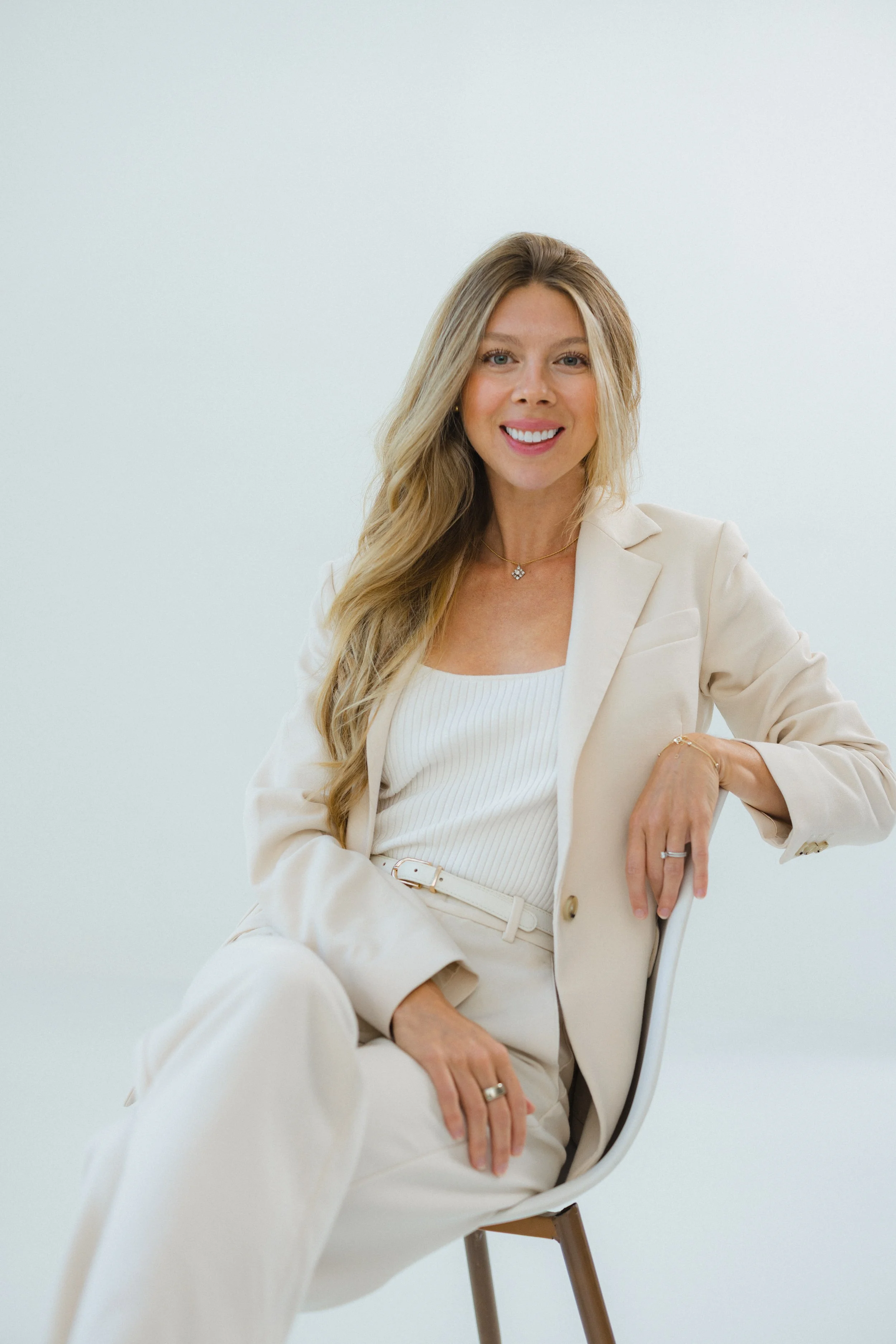 A woman with long blonde hair, wearing a cream blazer and matching pants, sitting on a chair with a white background.