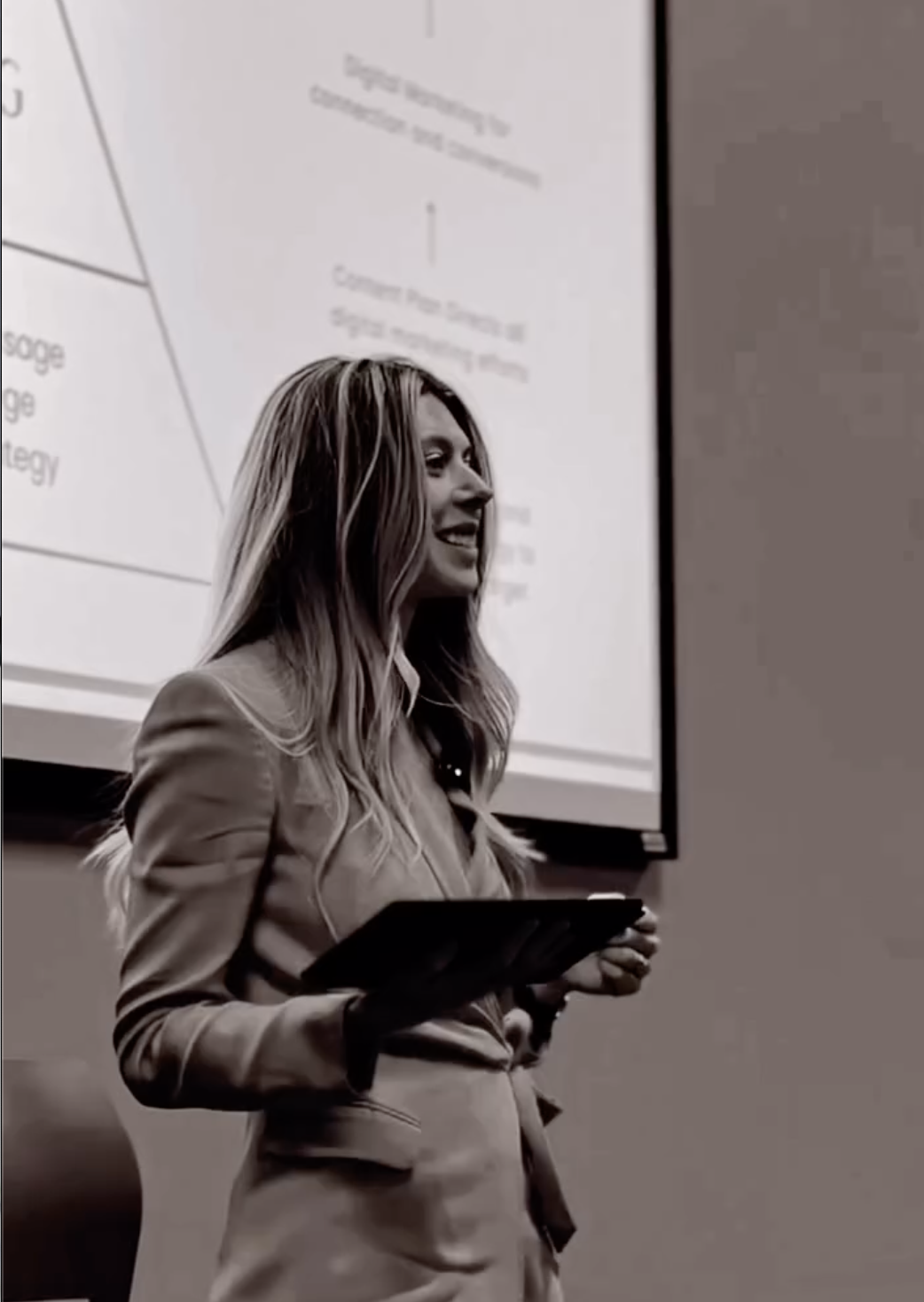 A woman holding a tablet in front of a presentation screen, smiling during a presentation or lecture.