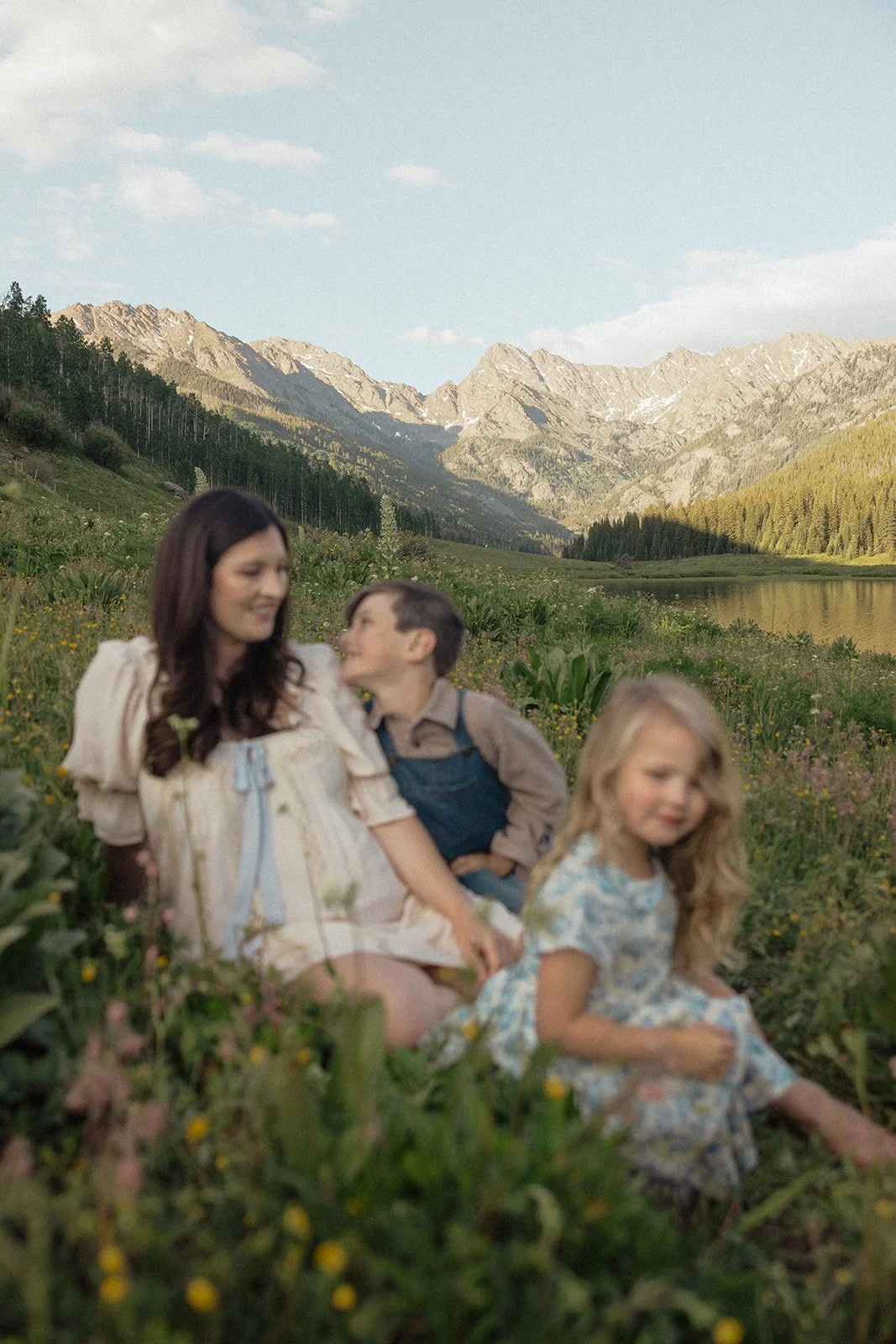 A woman and two children sitting in a meadow with mountains and a lake in the background.