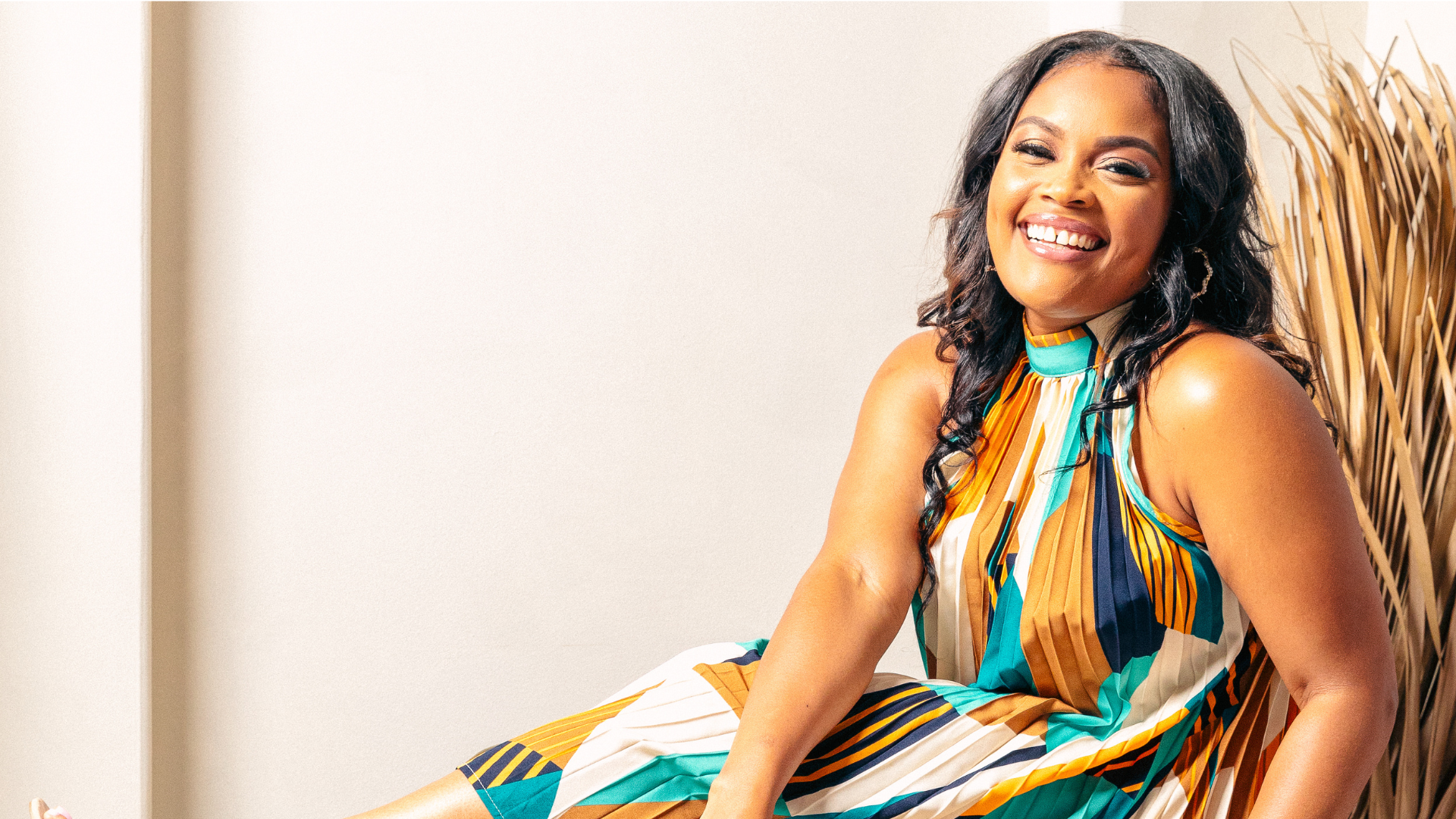 A woman with long dark wavy hair, smiling, wearing a colorful, striped sleeveless dress with a high neckline, sitting against a neutral wall and decorative dried plants in the background.