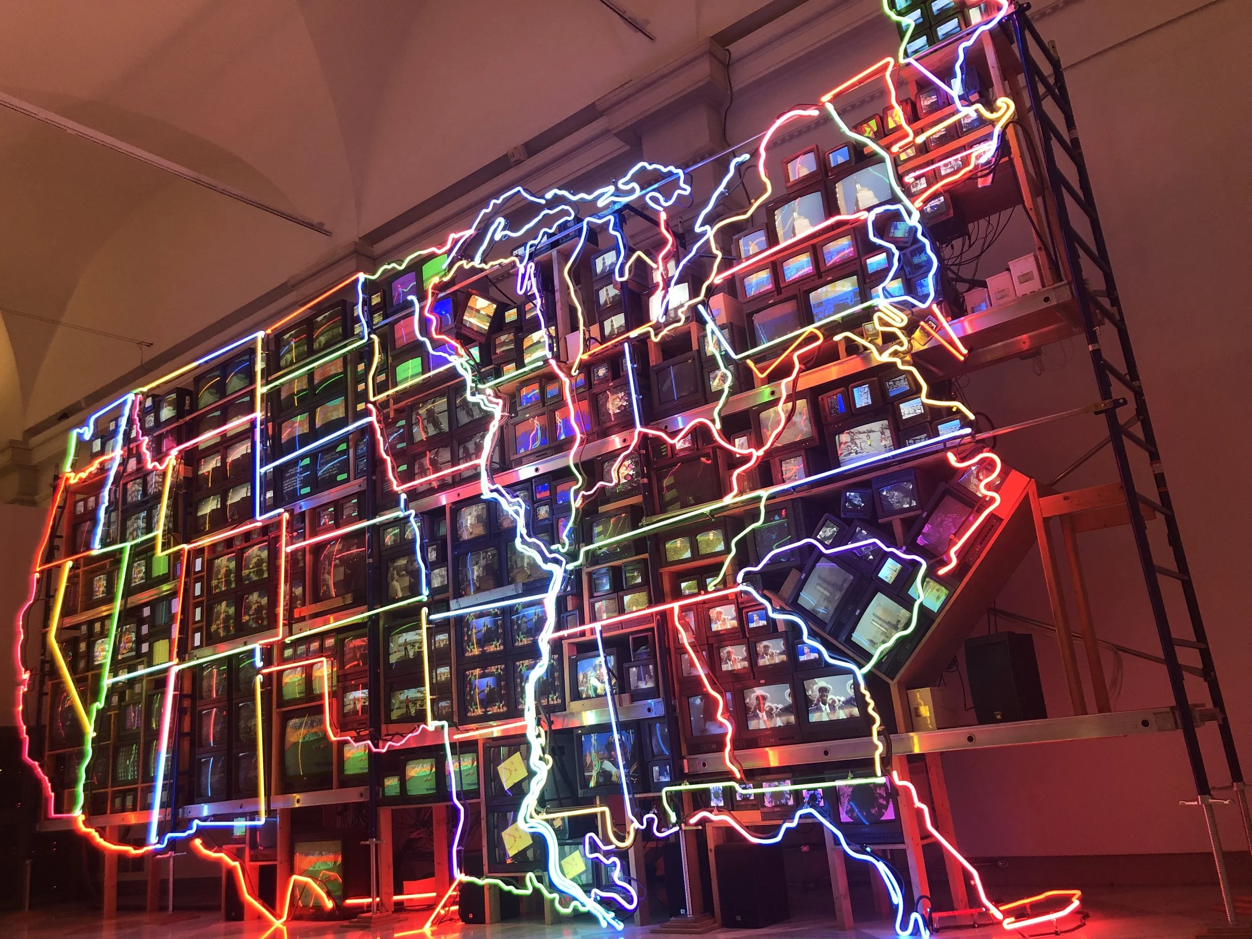 Neon outline of the map of North America with bright colorful neon lights, mounted on a wall with shelves holding vintage computer monitors.