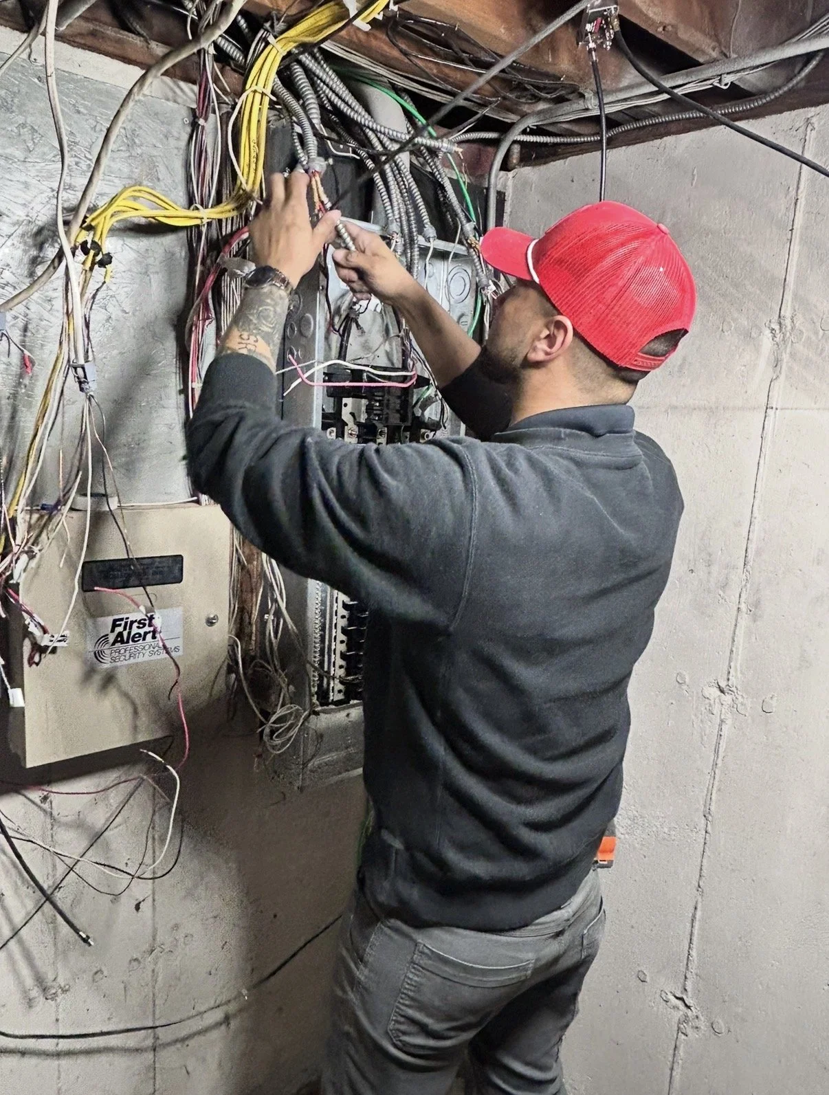 Man working on electrical wiring in an unfinished basement, wearing a red baseball cap, black jacket, and gray pants. He is fixing or installing electrical wires next to a fuse box and security system panel, surrounded by numerous wires and cables on a concrete wall.