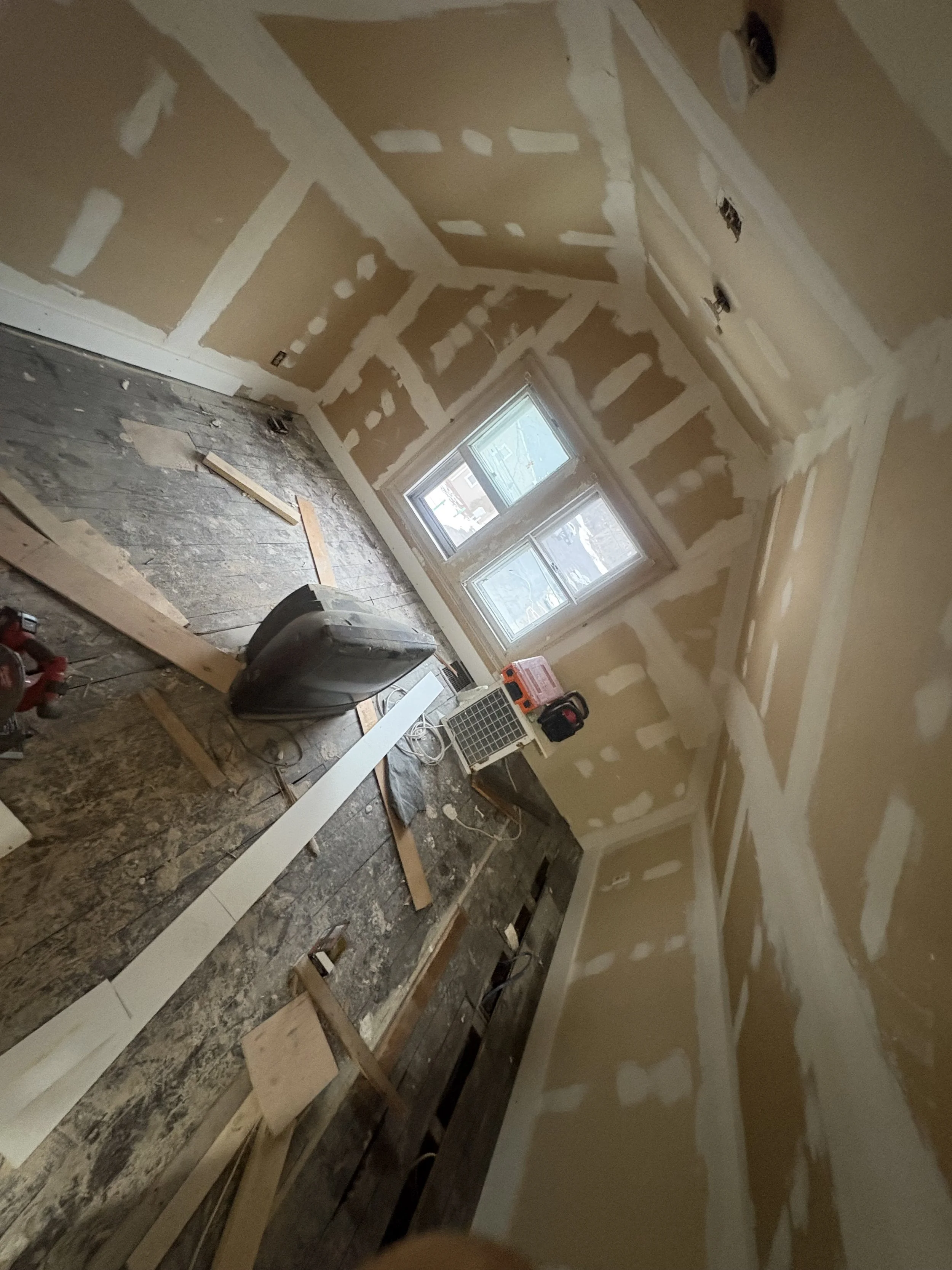 Interior of a room under renovation, with drywall installed on the walls and ceiling, and construction tools and materials on the floor near a window.