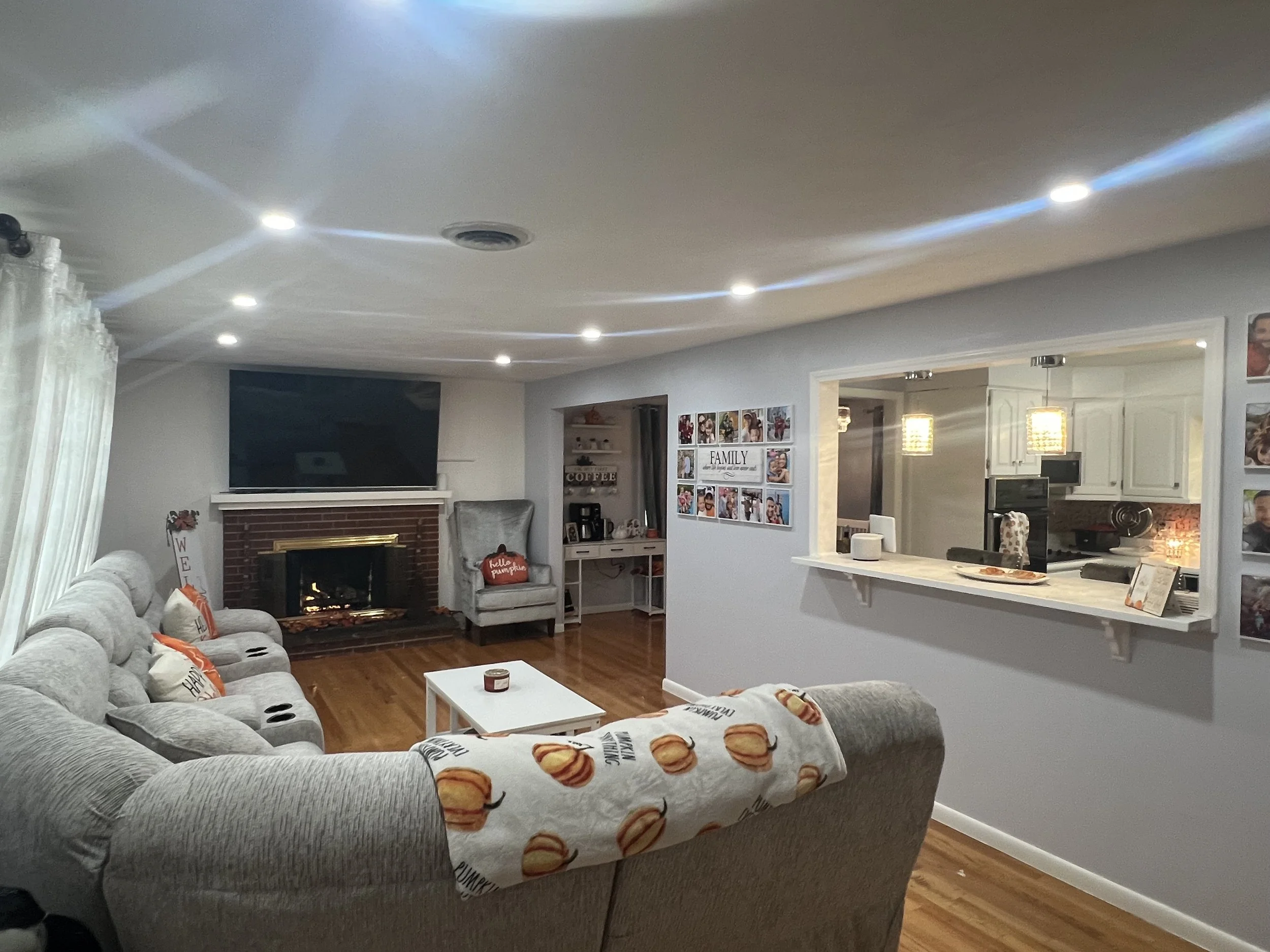 Living room with gray sectional sofa decorated with pumpkin-themed pillows, a white coffee table, a media console with a large flat-screen TV, a fireplace, and framed family photos on the wall. The ceiling has built-in lighting and a vent, with a kitchen visible in the background featuring white cabinets and pendant lights.