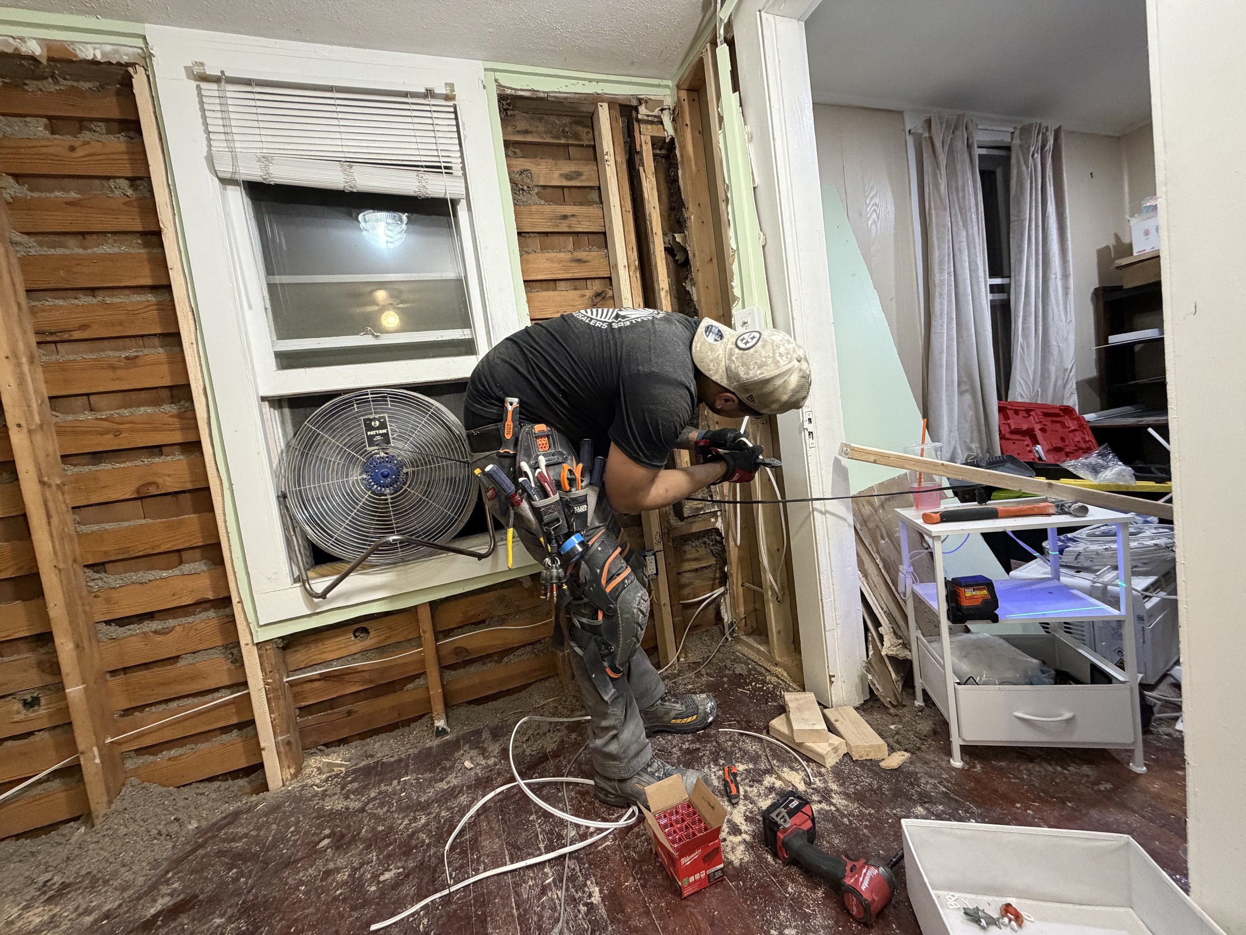 A man working on a home renovation project, kneeling and using tools to work on a wall that is partially torn down. The room has exposed wooden studs, a window with blinds, a small fan, and various tools and equipment scattered around.