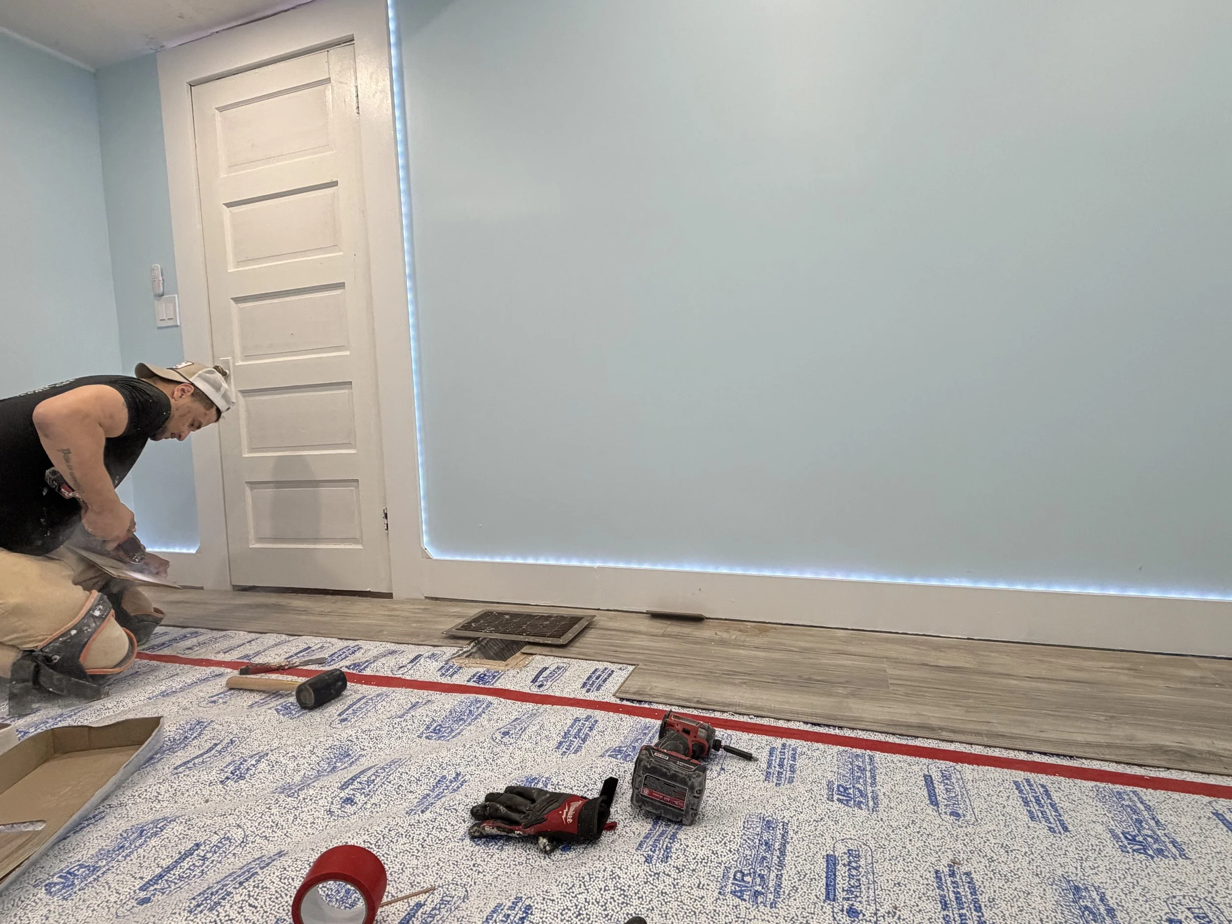 A man working on a home renovation project in a room with light blue walls. Tools and materials are scattered on the floor, including drills, a level, and protective tape. The door is white with horizontal panels, and there is a strip of LED lighting near the ceiling.
