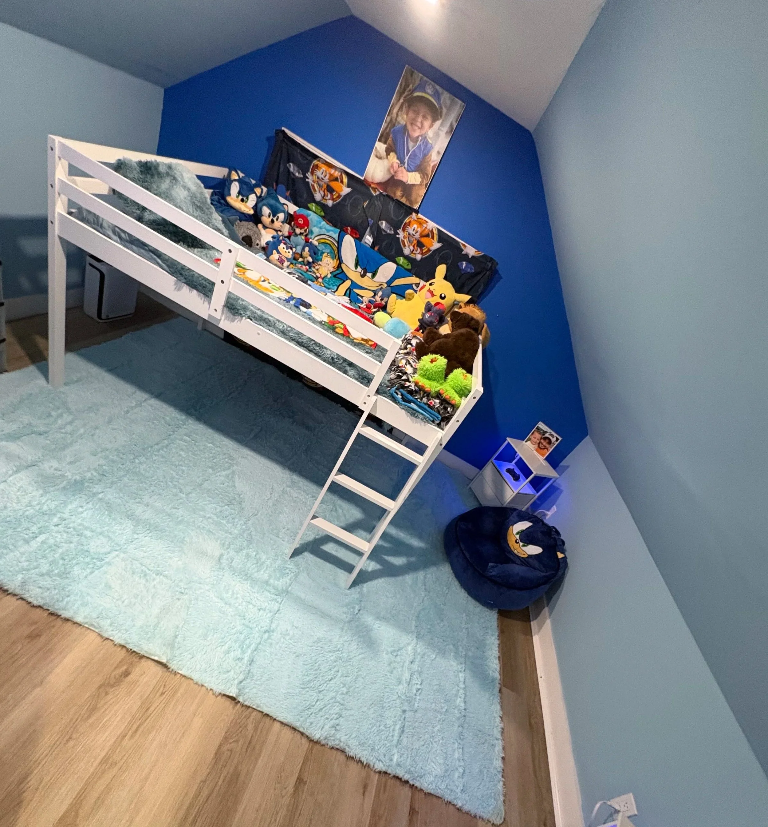 A bedroom with a light blue rug, a white loft bed with stuffed animals, including Sonic the Hedgehog, Pikachu, and other plush toys. The back wall is painted dark blue, with a large family photo hanging on it. There is a small white nightstand with a photo frame and a black backpack on the floor beside it.