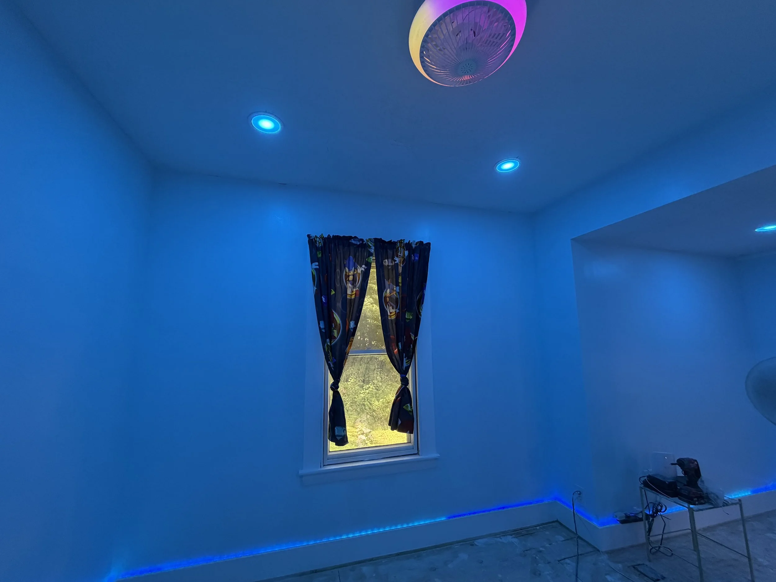A room with blue walls and ceiling, blue recessed lighting, a window with curtains featuring cartoon characters, and a ceiling fan with pink and yellow lighting.