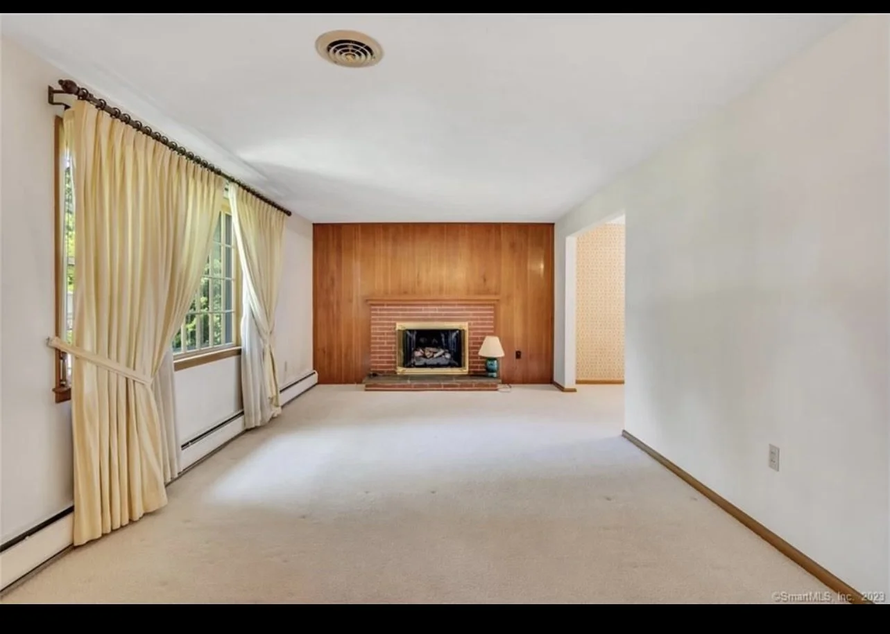 Empty living room with beige carpet, cream-colored curtains, a large window on the left, a brick fireplace with a wooden panel behind it, and a small green table lamp next to the fireplace.