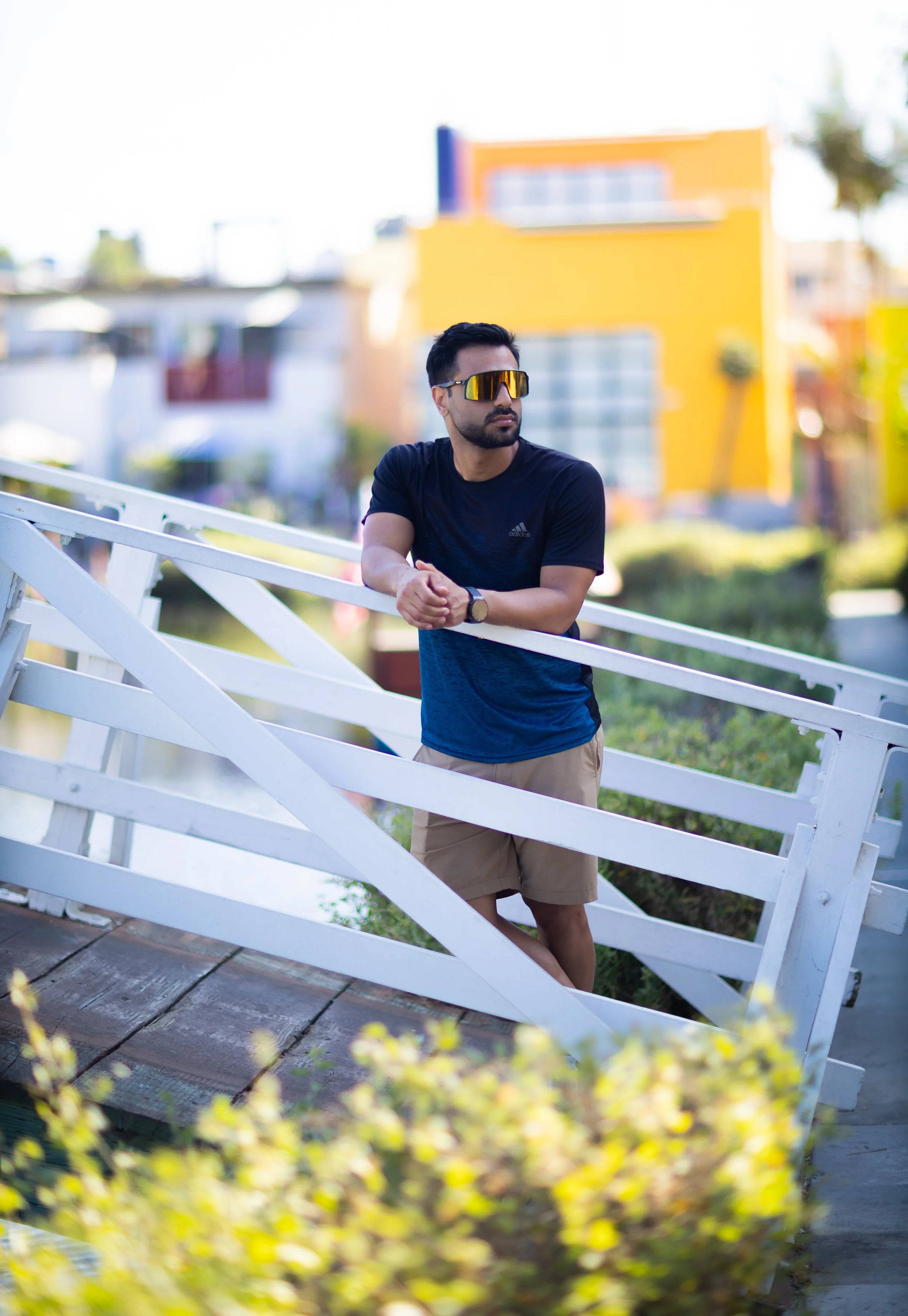 Man wearing sunglasses and a black Adidas shirt leaning on a white wooden fence on a sunny day, with blurred colorful buildings in the background.
