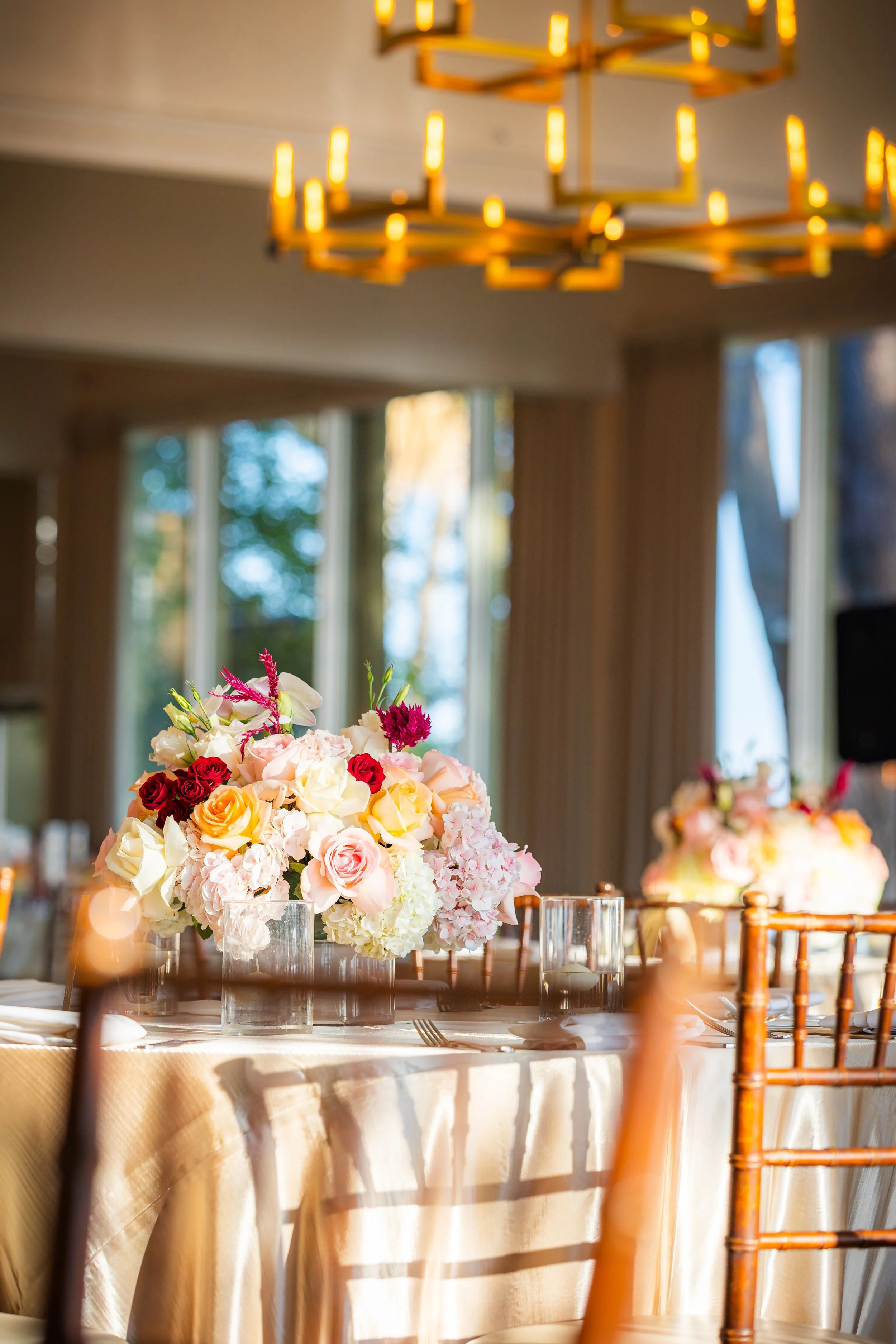 A tablescape with a bouquet of pink, white, red, yellow, and purple flowers as a centerpiece, set with glassware, white napkins, and a beige tablecloth, on a wooden table in a room with large windows and a gold-colored chandelier.