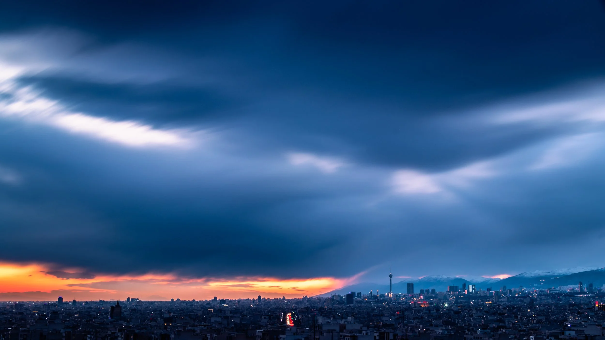 A city skyline at sunset with colorful clouds in the sky and buildings silhouetted against the horizon.