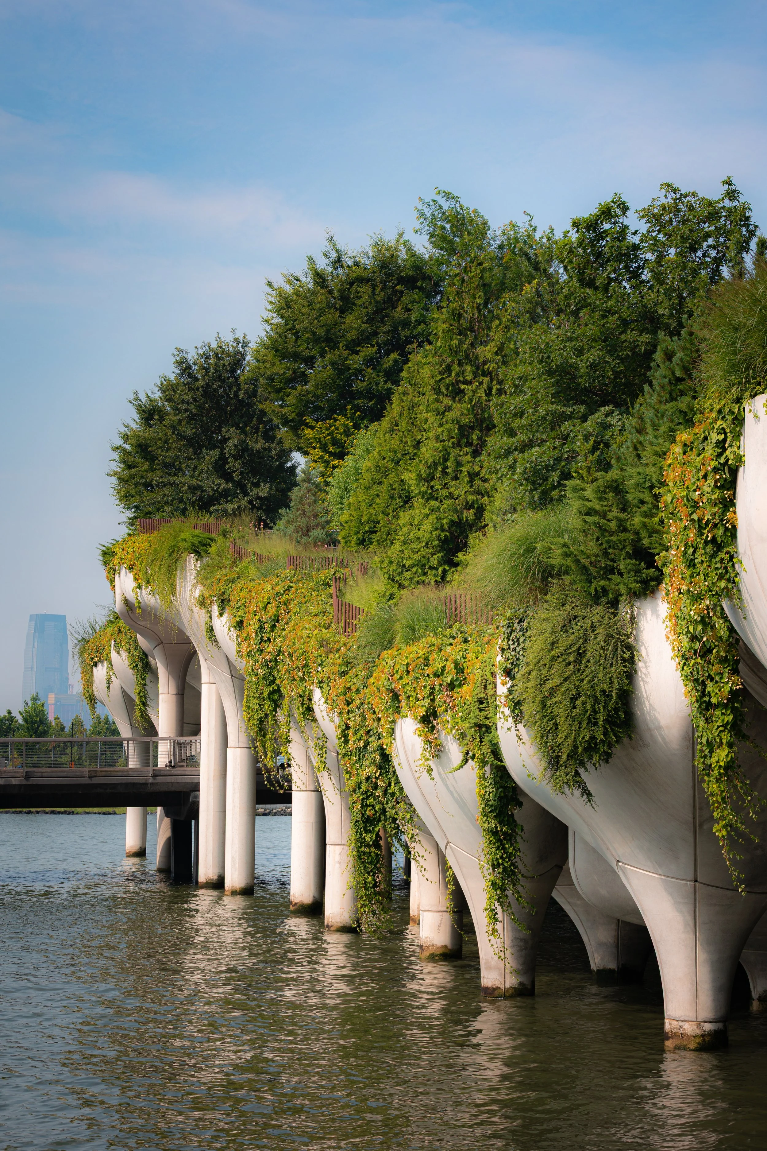A modern architectural structure with white supports and a lush green roof, situated along a body of water, with a city skyline in the background under a blue sky.
