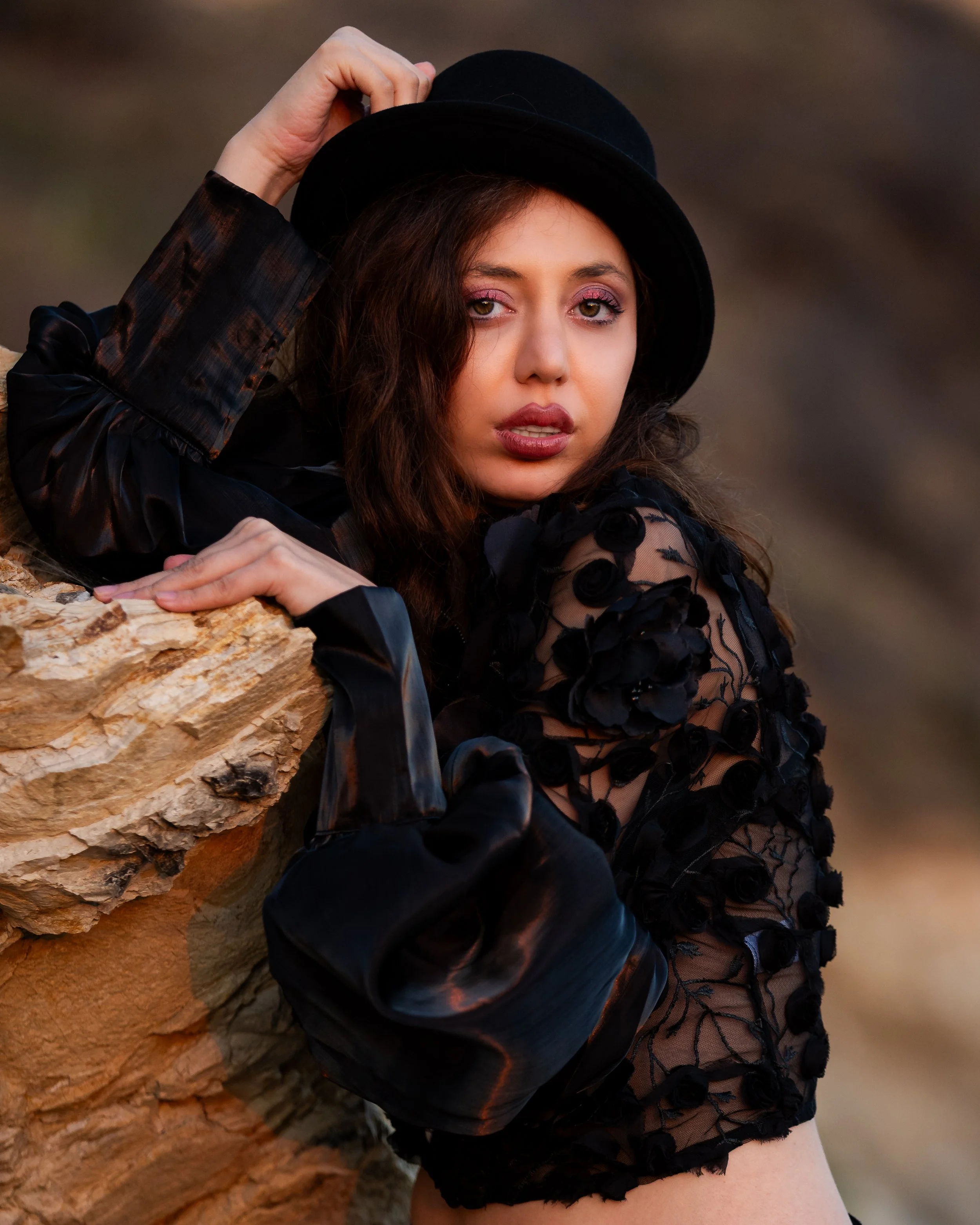 A woman leaning against a rocky surface, wearing a black hat and black clothing with floral overlay, looking directly at the camera with a neutral expression.