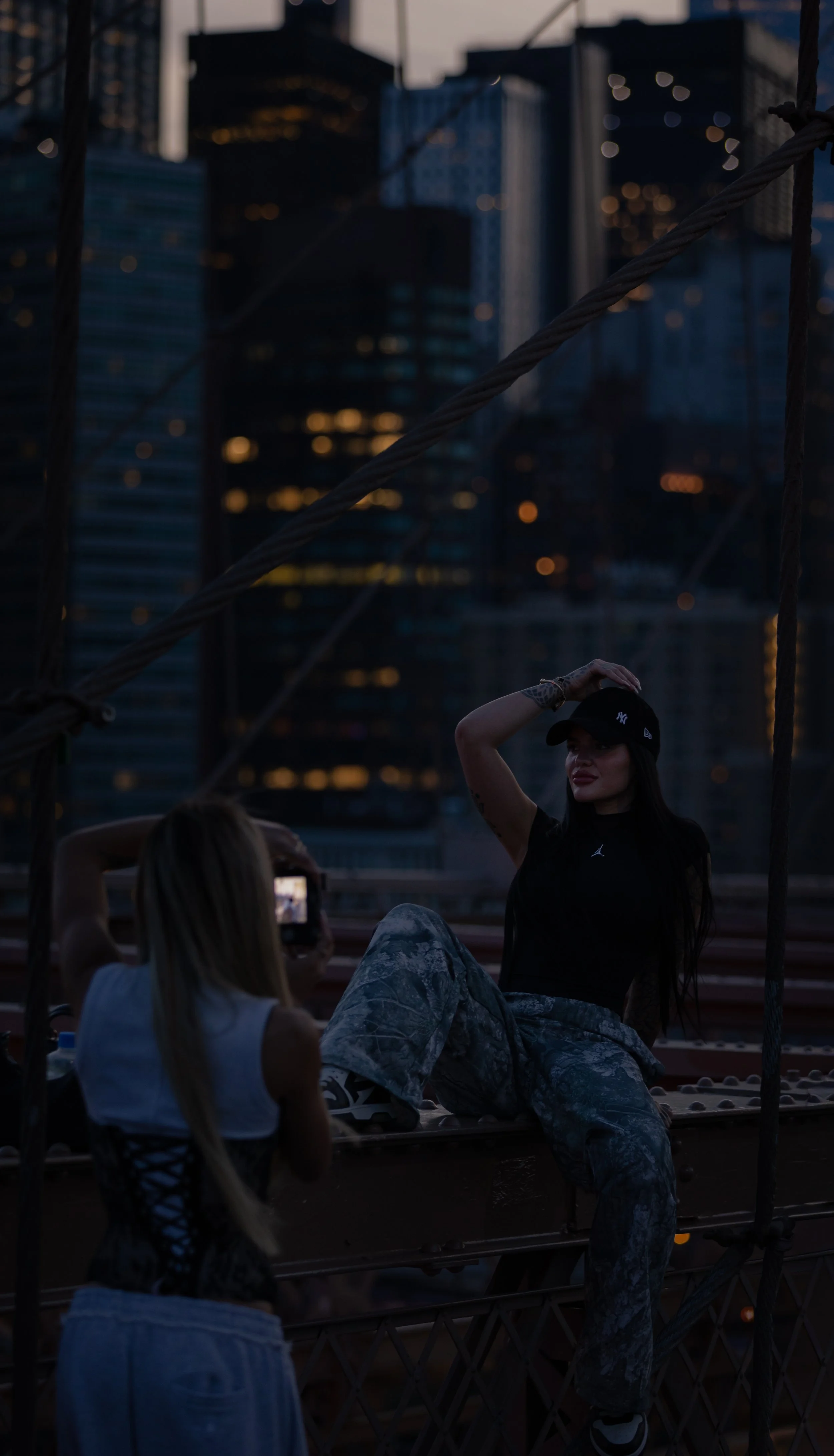 A woman with long dark hair wearing a black cap and black shirt with a white logo, sitting on a bridge at dusk, surrounded by city skyscrapers with illuminated windows, while a person in front of her takes a photo.