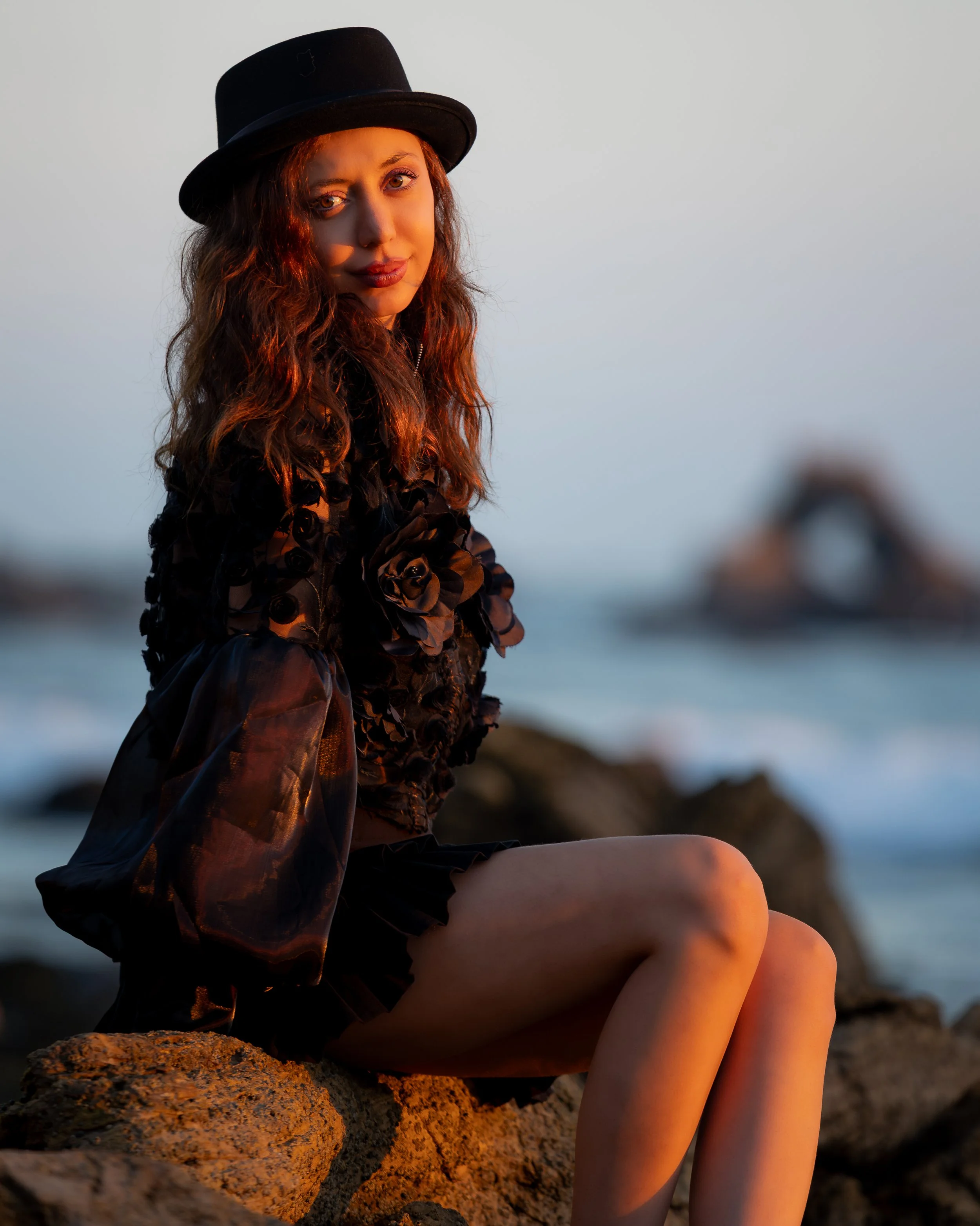 A woman with long, curly hair wearing a black hat and a black dress with roses, sitting on rocks at sunset near the water.