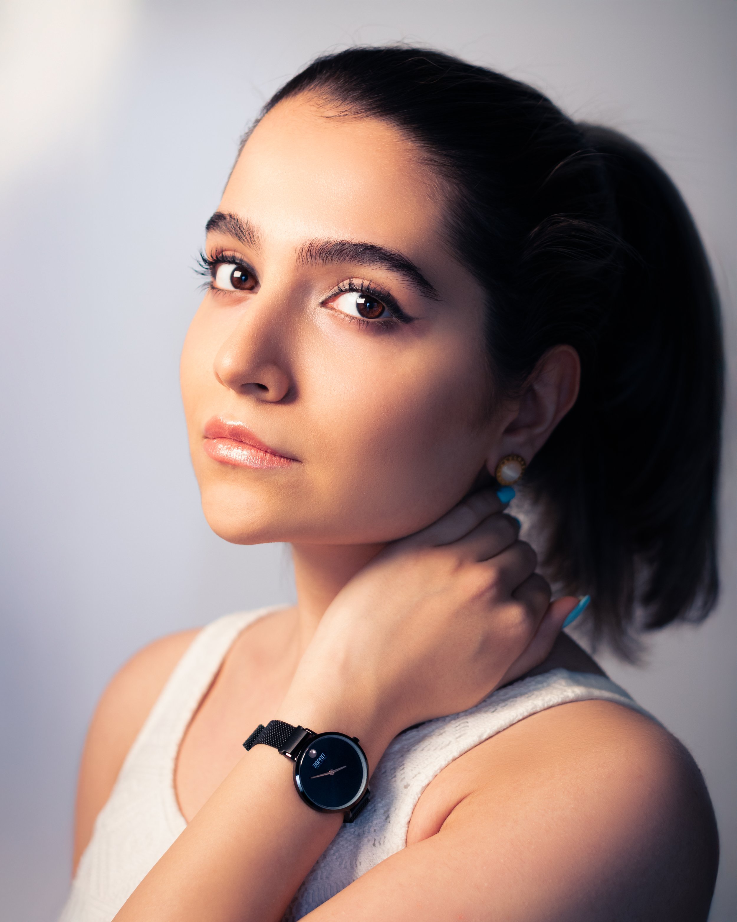 Close-up of a young woman with dark hair, looking away from the camera, wearing a sleeveless white top, and a black wristwatch with a round face on her left wrist.