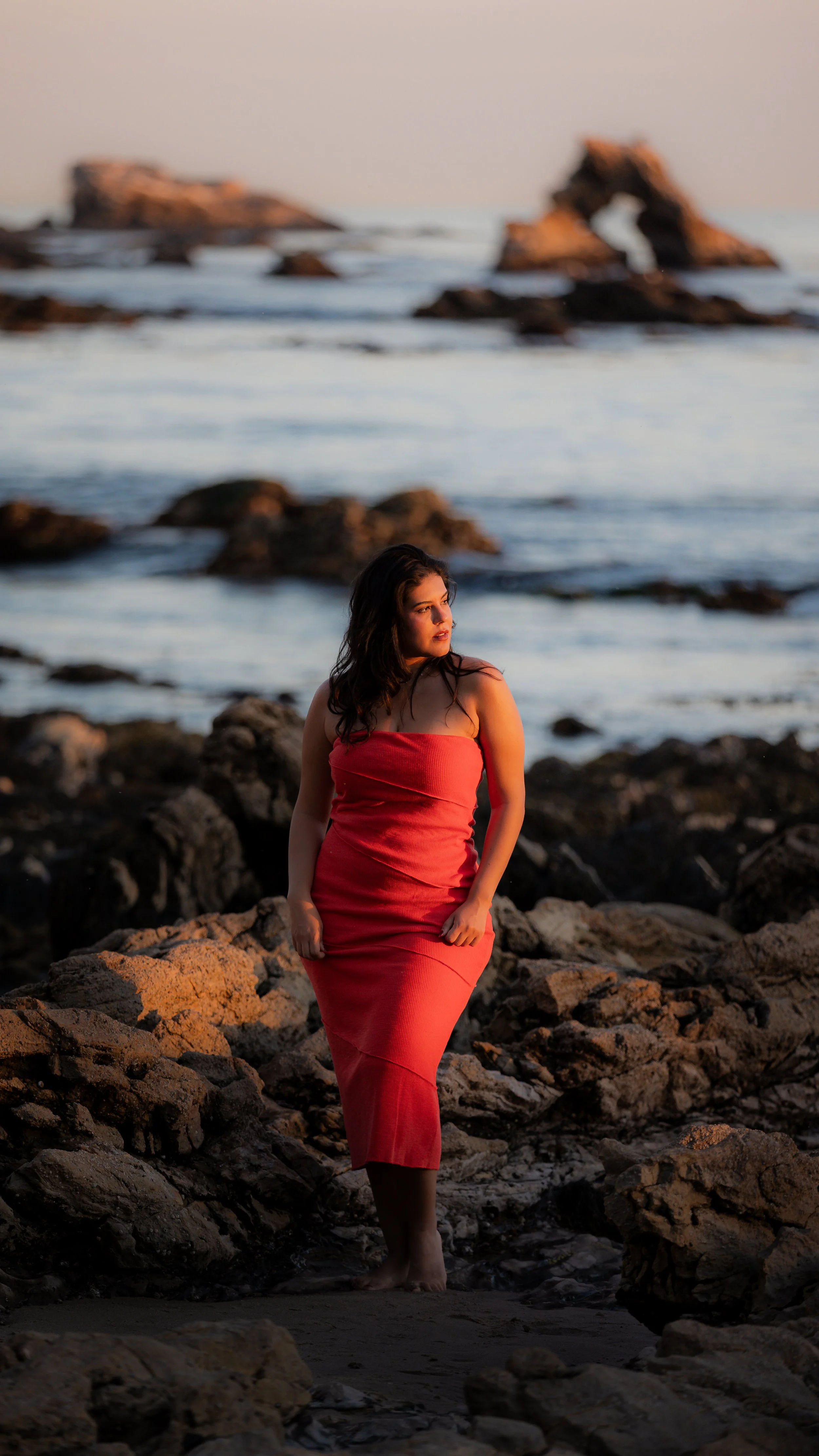 A woman in a red dress standing on rocks by the ocean, with a rocky arch and formations in the background at sunset.