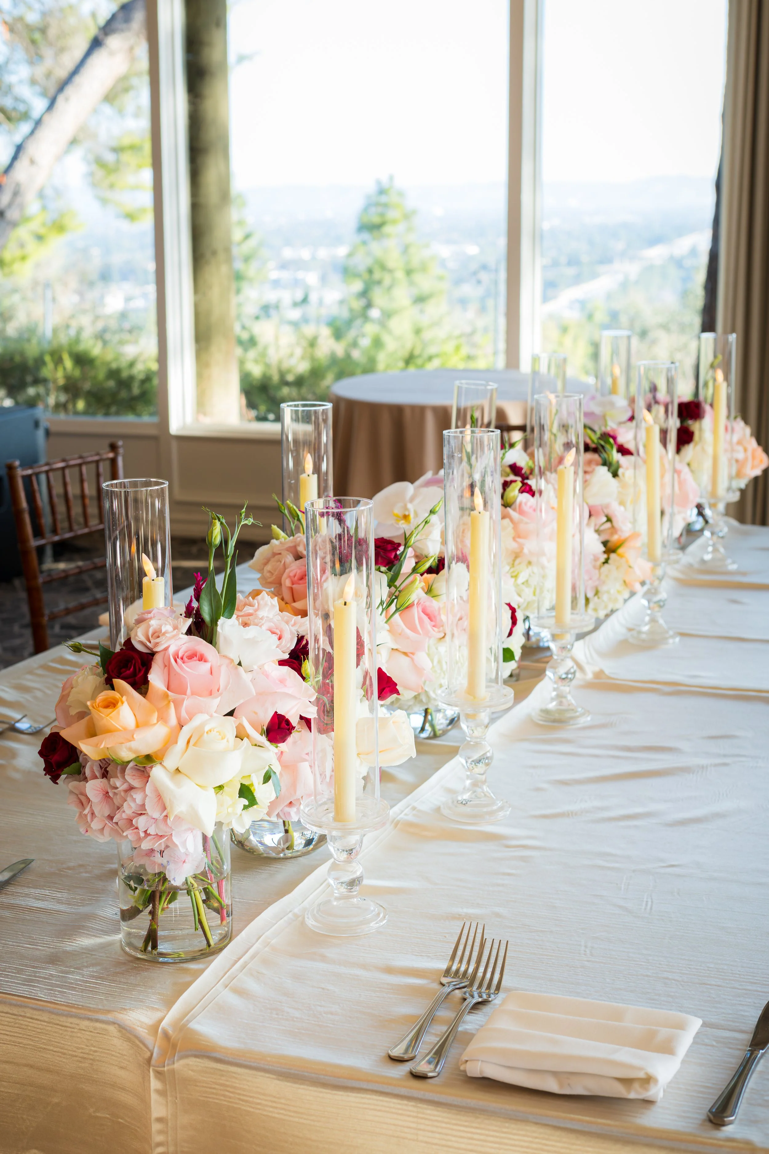 A decorated banquet table set for a formal event with floral centerpieces and candles, with large windows overlooking a scenic outdoor landscape.