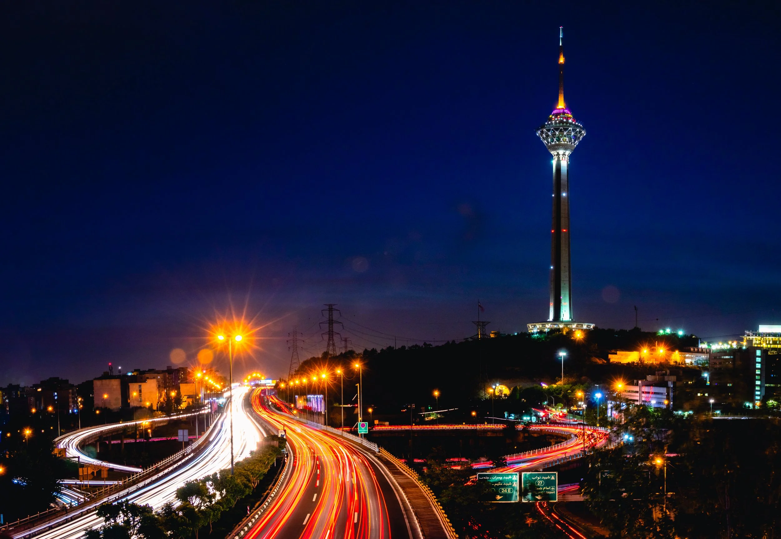 Night view of a city with a tall illuminated tower, city lights, and light trails from moving vehicles on a highway.