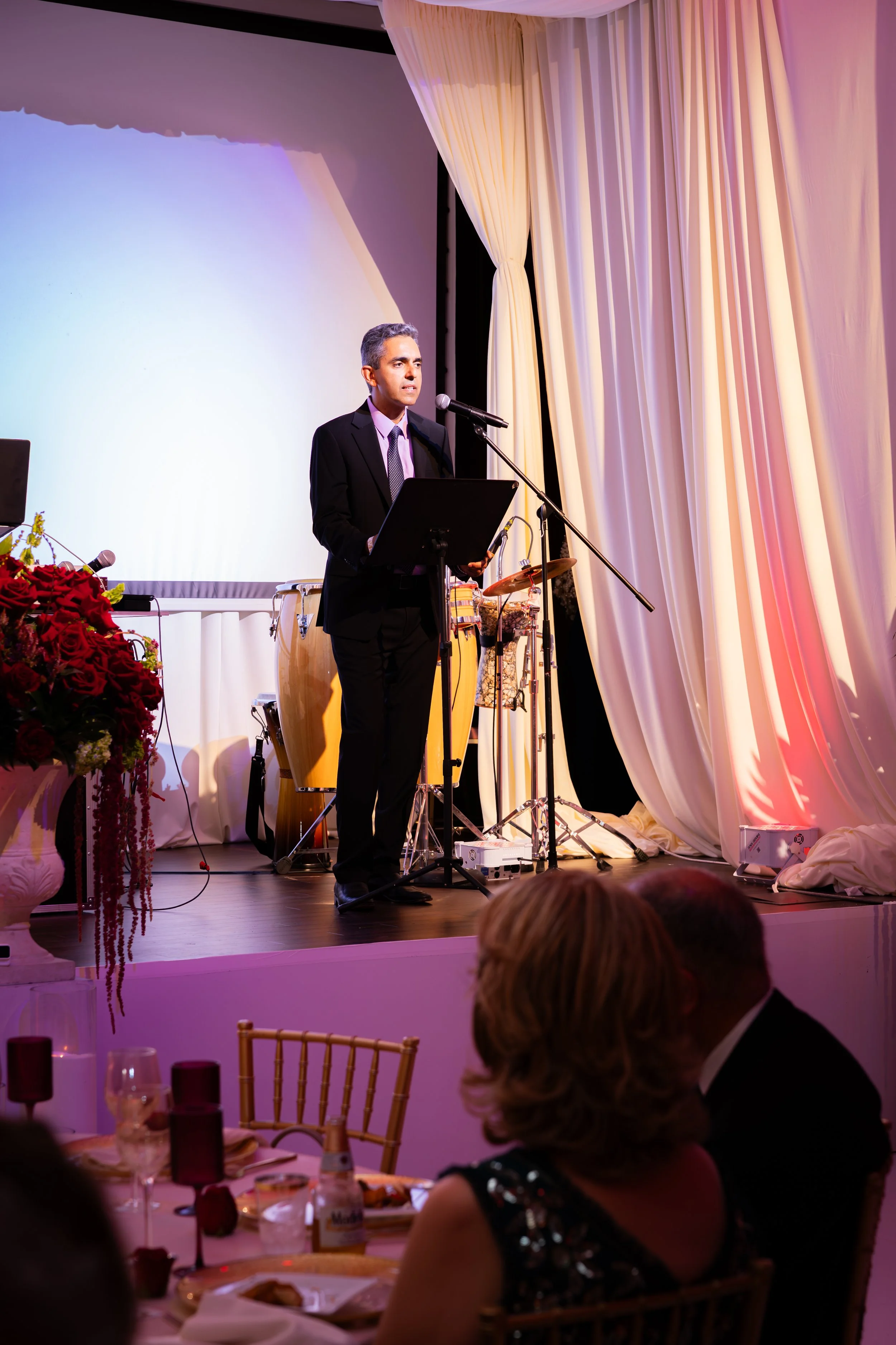 A man in a suit standing at a podium giving a speech with musical instruments behind him on stage, decorated with large white curtains and pink lighting, during a formal event.