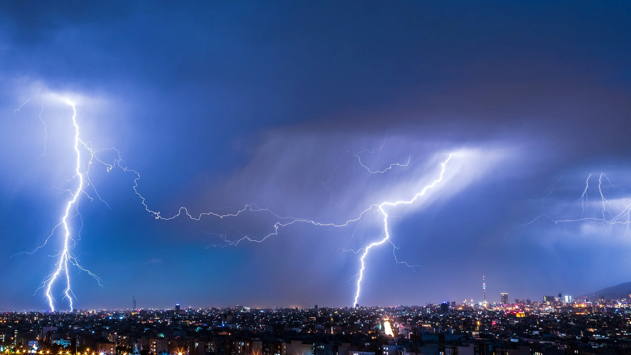 Multiple lightning strikes illuminate a dark, cloudy sky over a cityscape at night.
