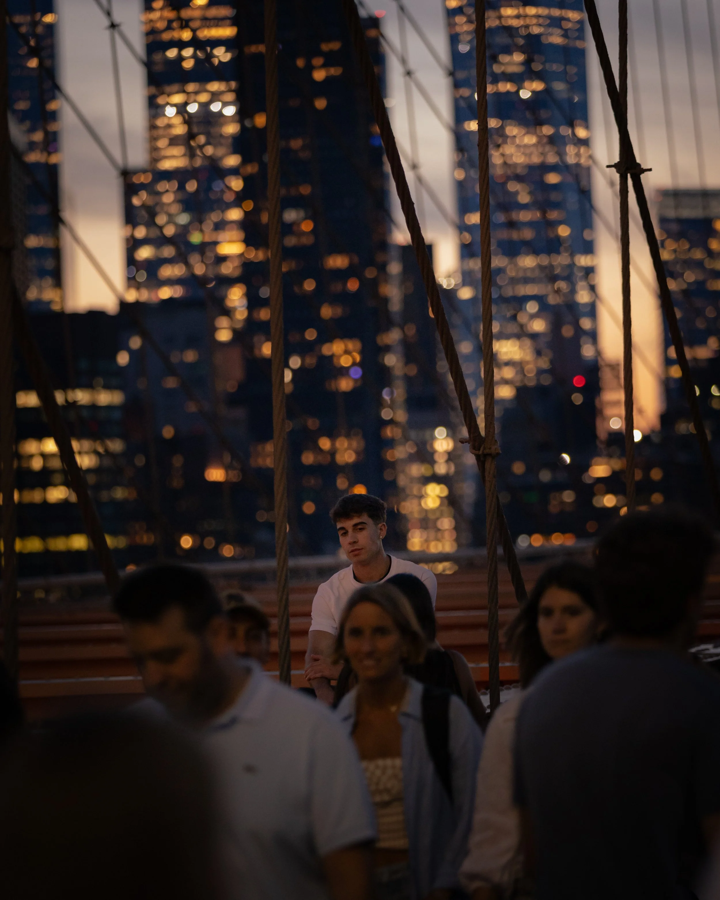 A group of people sitting outdoors during sunset, with a city skyline in the background. The focus is on a young man with dark hair, wearing a white shirt, sitting among others who are slightly out of focus.