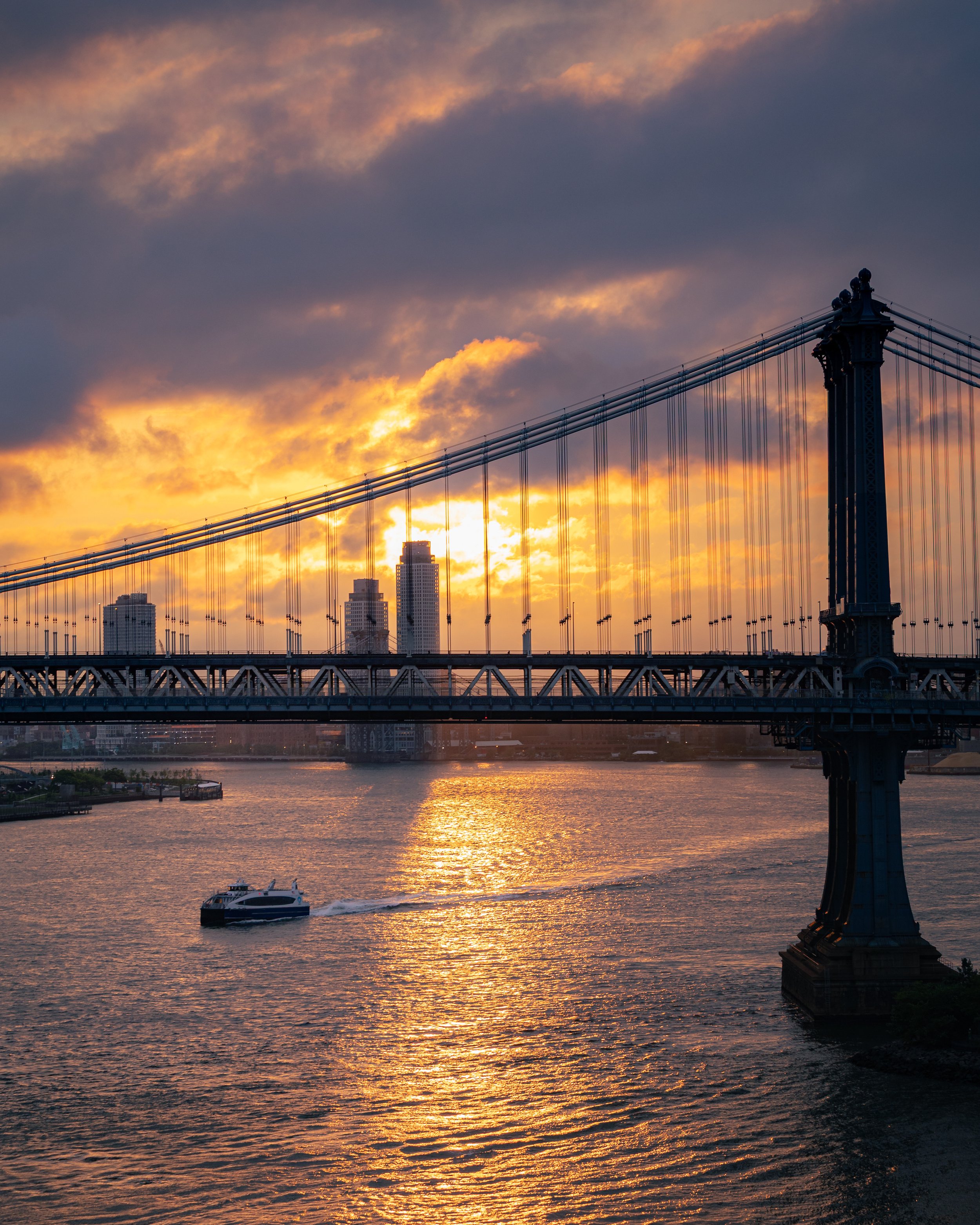 Sunset over a river with a boat, with the Brooklyn Bridge in the foreground and city buildings in the background.