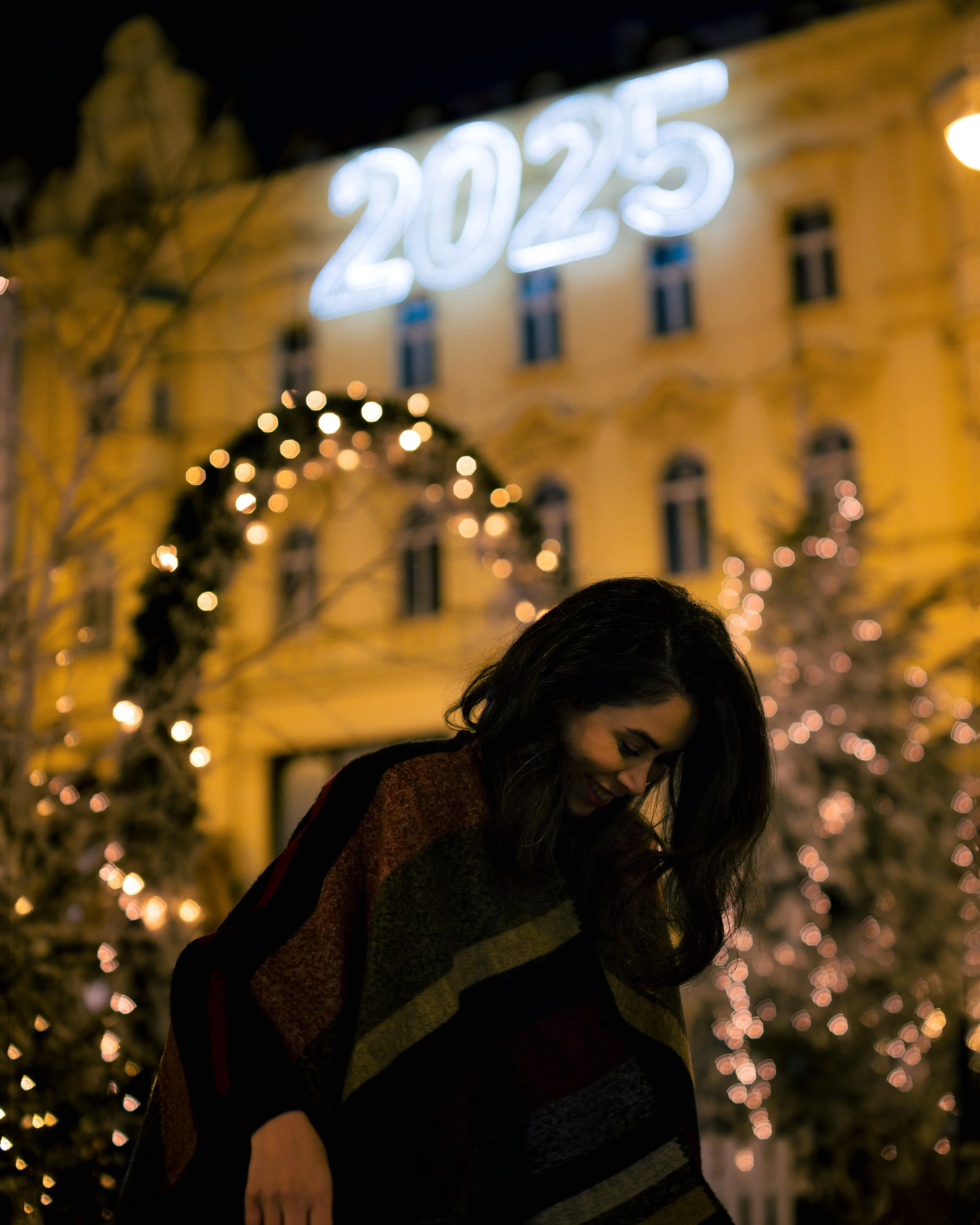 Night scene with a smiling woman in front of a decorated Christmas tree with blurred warm lights and a building with a sign reading '2025' above.