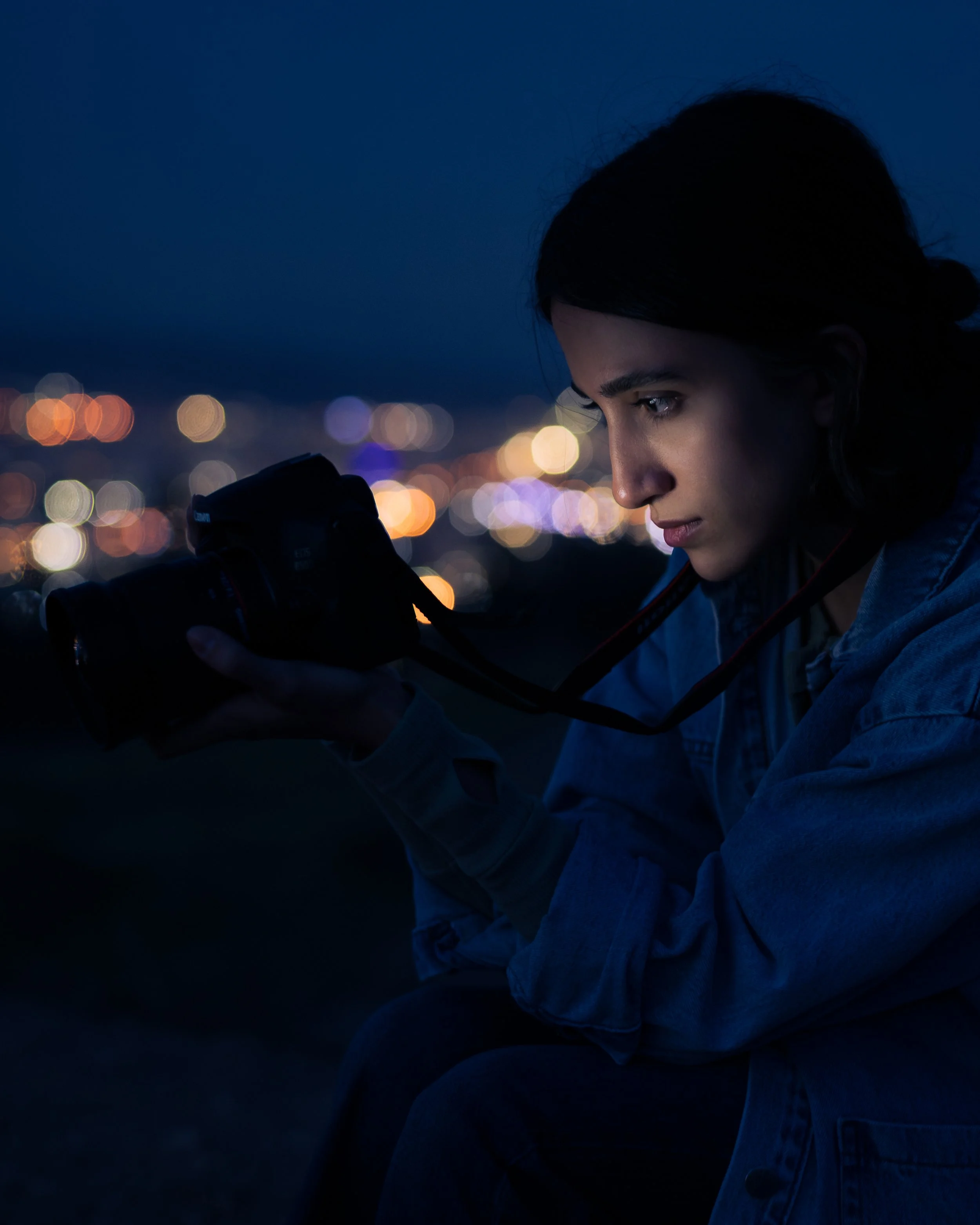 A young woman with dark hair looking down at a camera in her hands during nighttime with blurred city lights in the background.