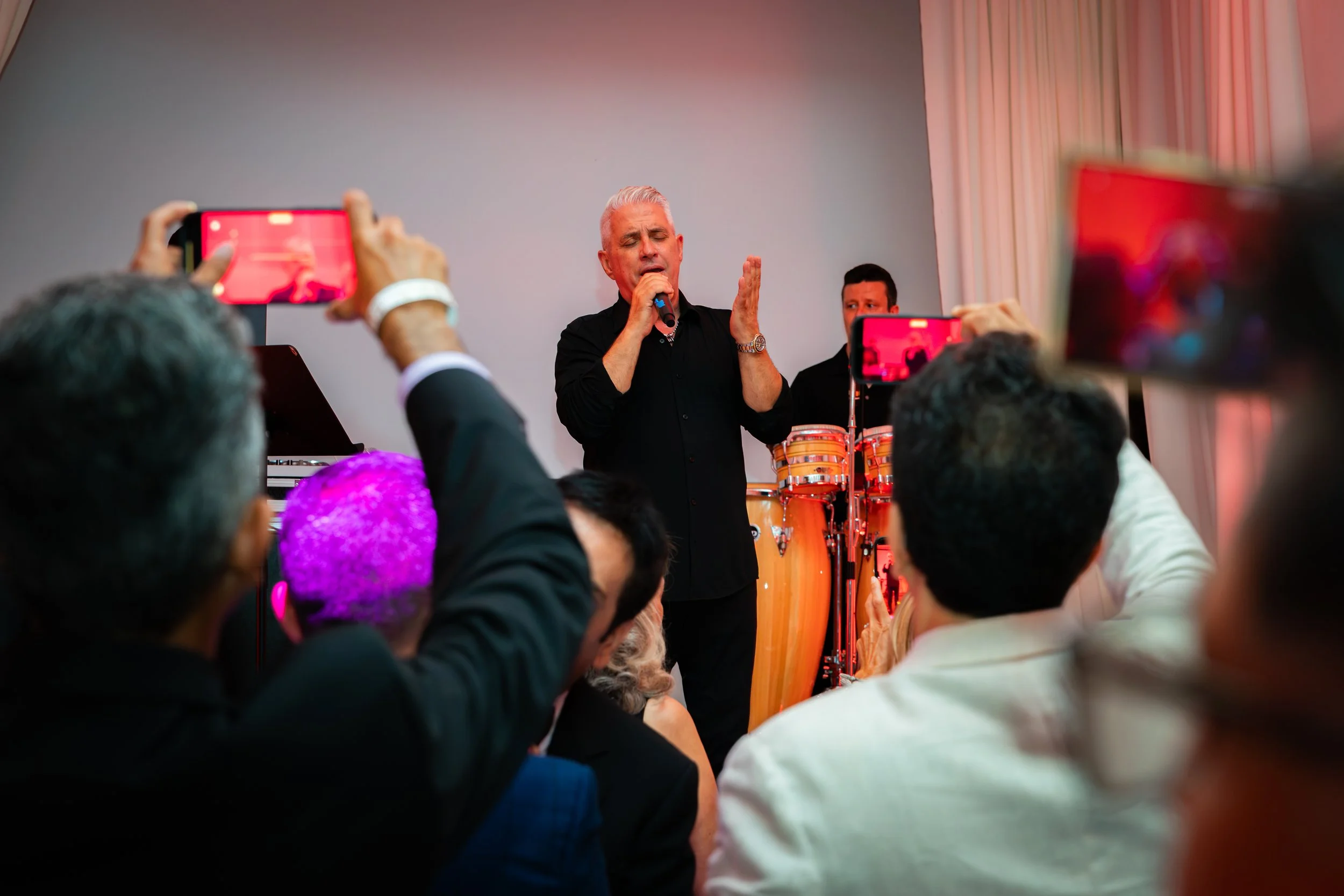 An older man with white hair singing into a microphone at a performance, with percussion instruments behind him, audience members taking photos, and diverse audience members in the foreground.