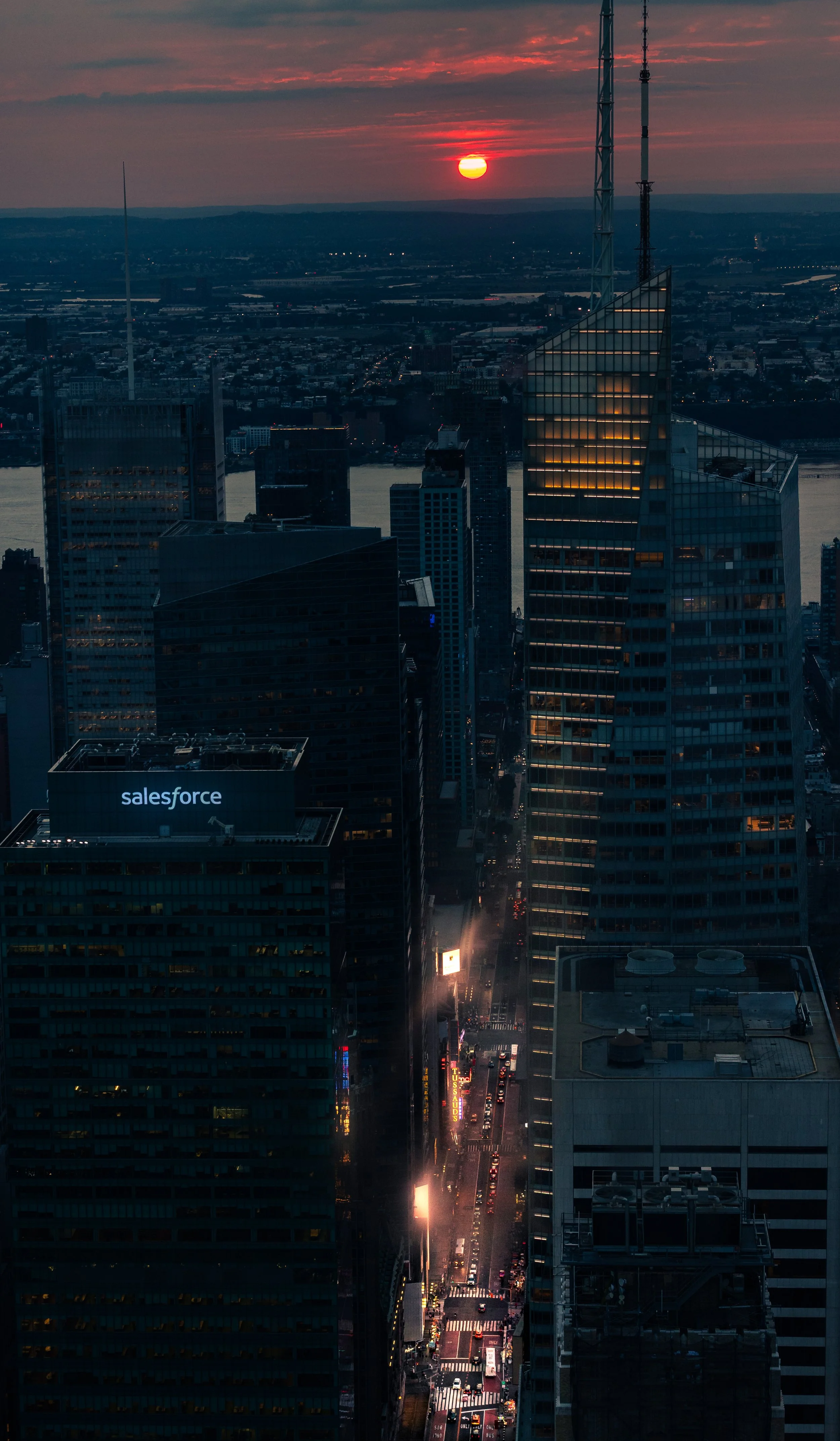 A cityscape at sunset with tall buildings, a river, and a busy street with cars and neon lights.