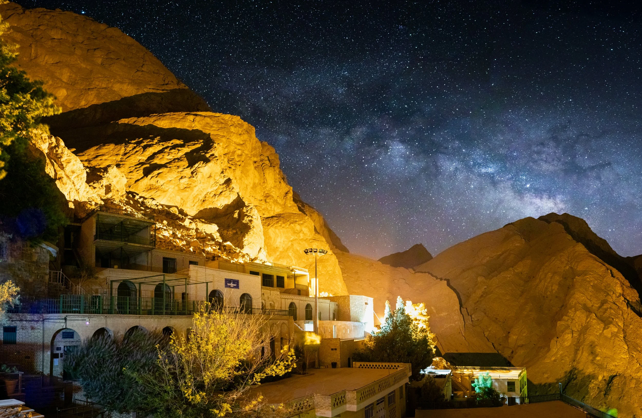 Mountain cliffs illuminated at night with a star-filled sky in the background, buildings and trees at the base.