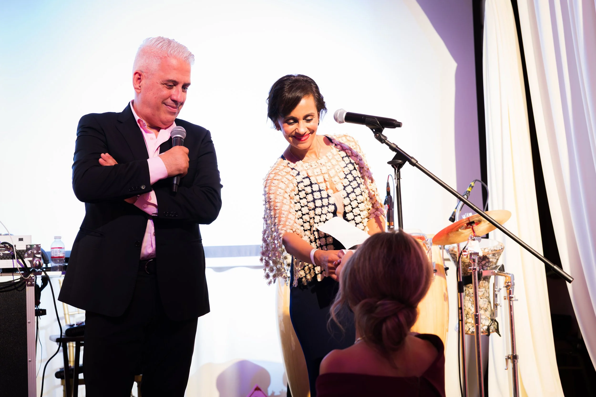 Two individuals on stage, one man in a dark suit and pink shirt holding a microphone, and one woman in a floral dress smiling and holding papers near a microphone stand, with musical instruments and equipment in the background at an event.