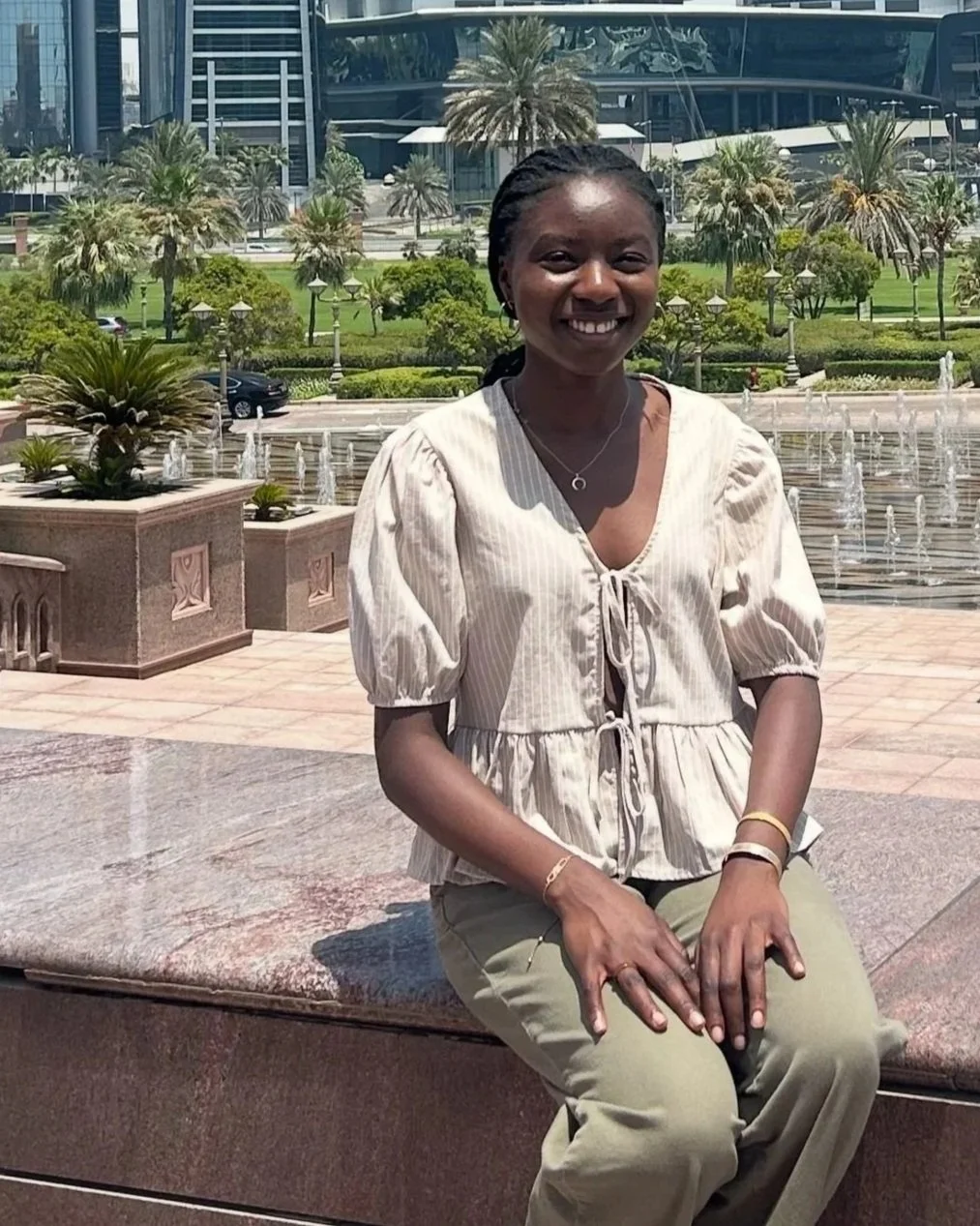 A woman sitting on a pink granite bench in front of a fountain in a landscaped urban park, with modern glass buildings and palm trees in the background, smiling at the camera.