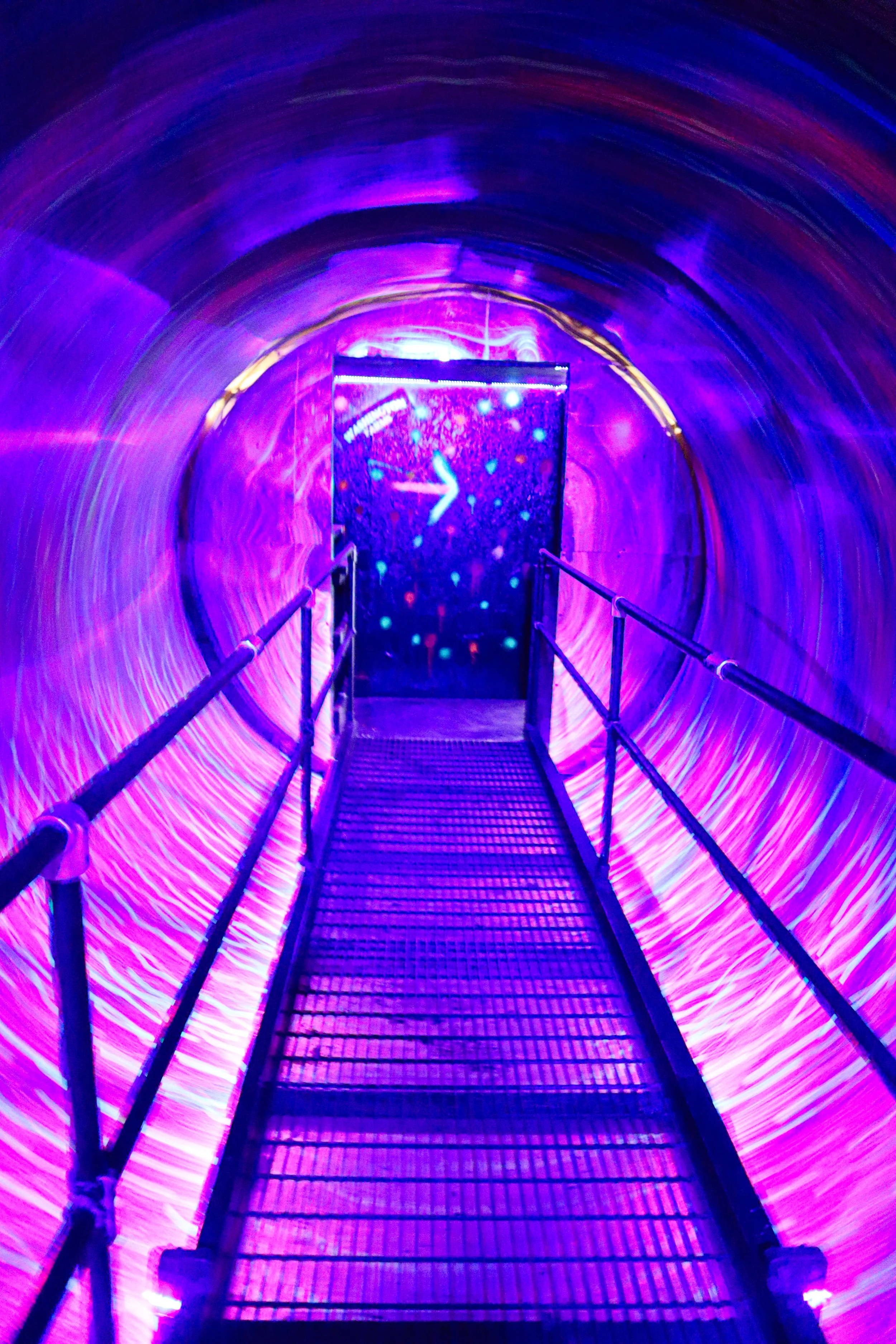 A glowing, illuminated tunnel with vibrant purple and pink light streaks, leading to a digital or LED screen at the end, with a metal walkway and railing.
