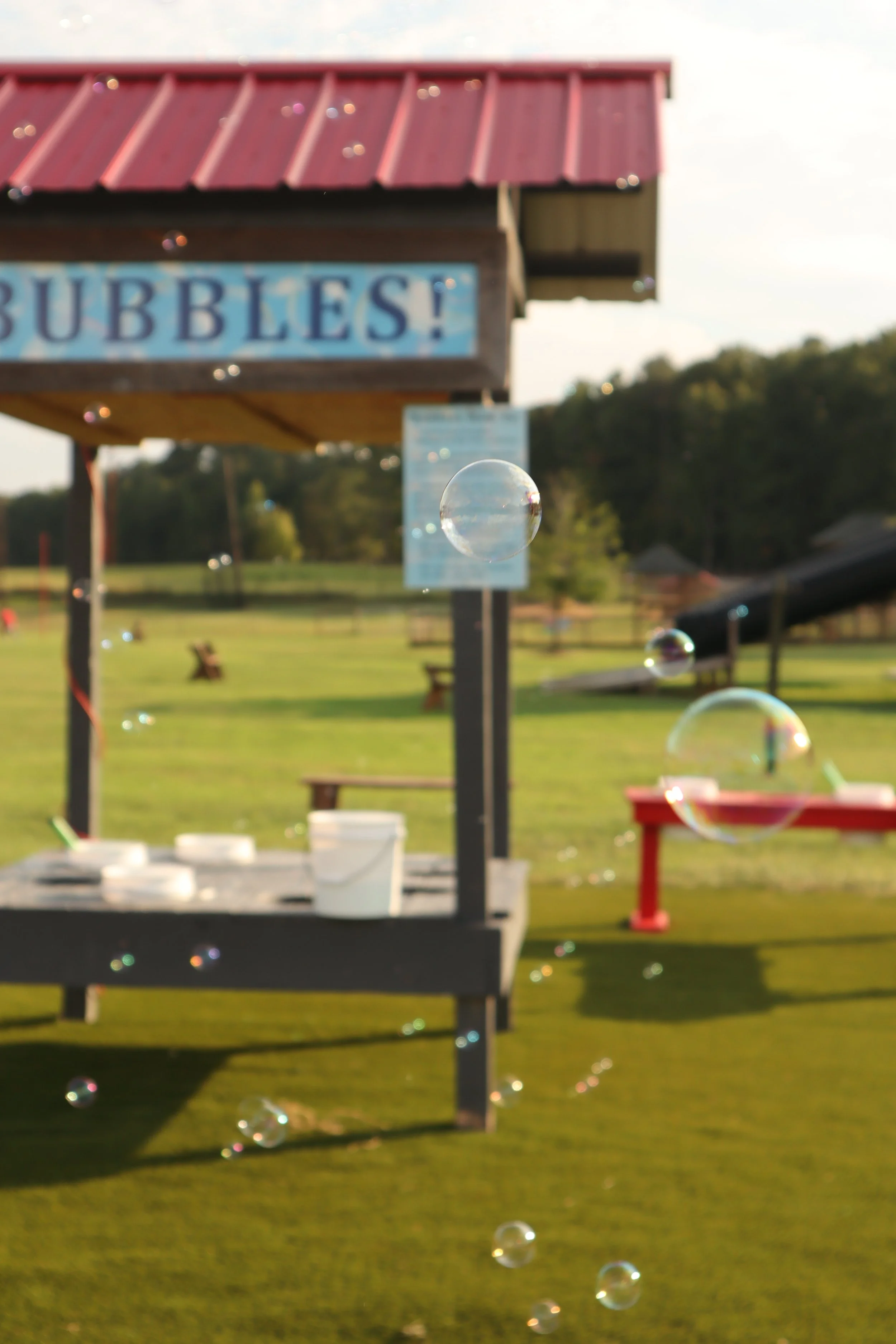 Bubbles floating in the air near a shed or booth with a sign that reads 'BUBBLES!'. The background includes green trees, grass, and outdoor furniture.