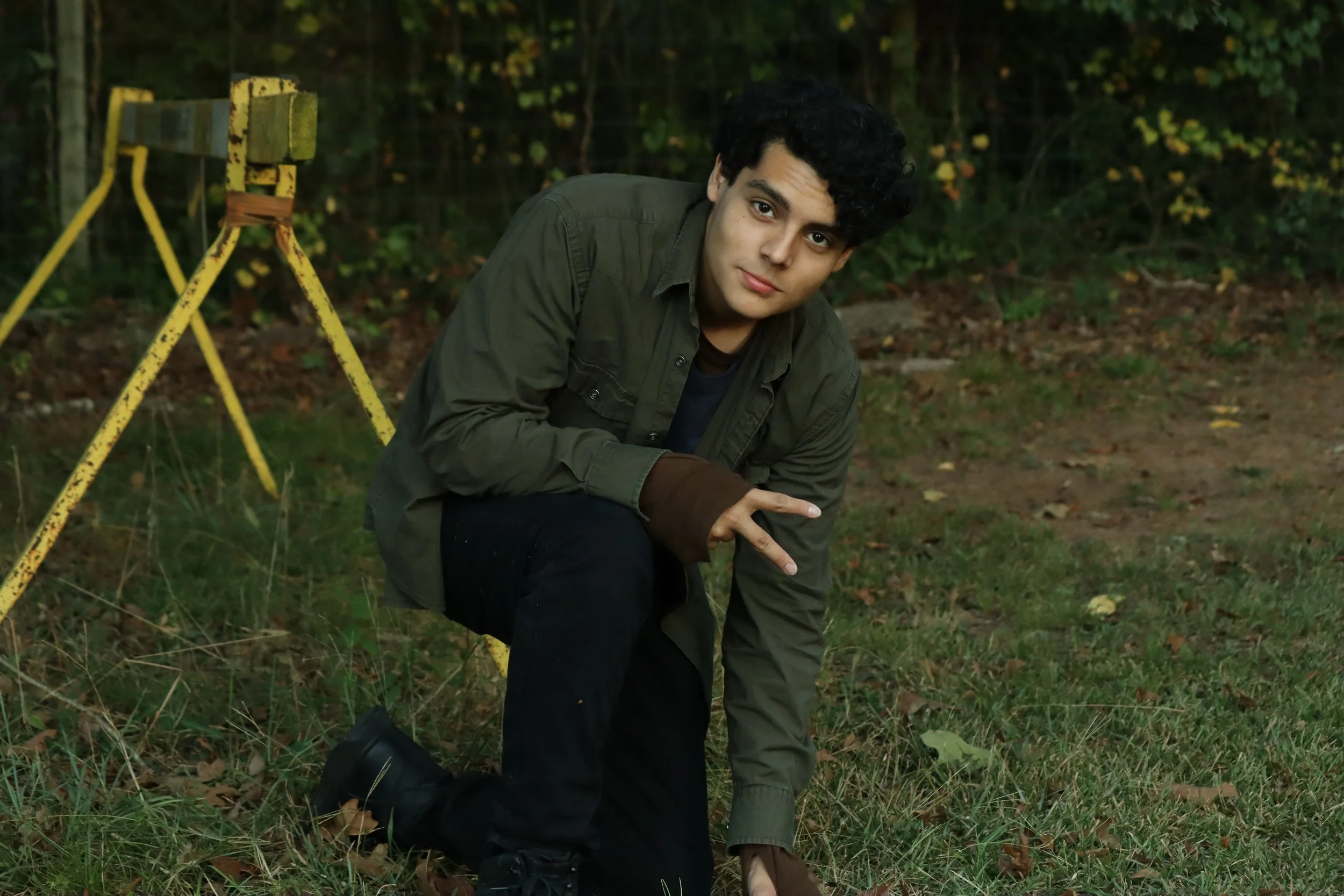 Young man crouching outdoors in a wooded area with yellow and green trees, wearing a dark green jacket, with an old yellow tripod or ladder in the background.