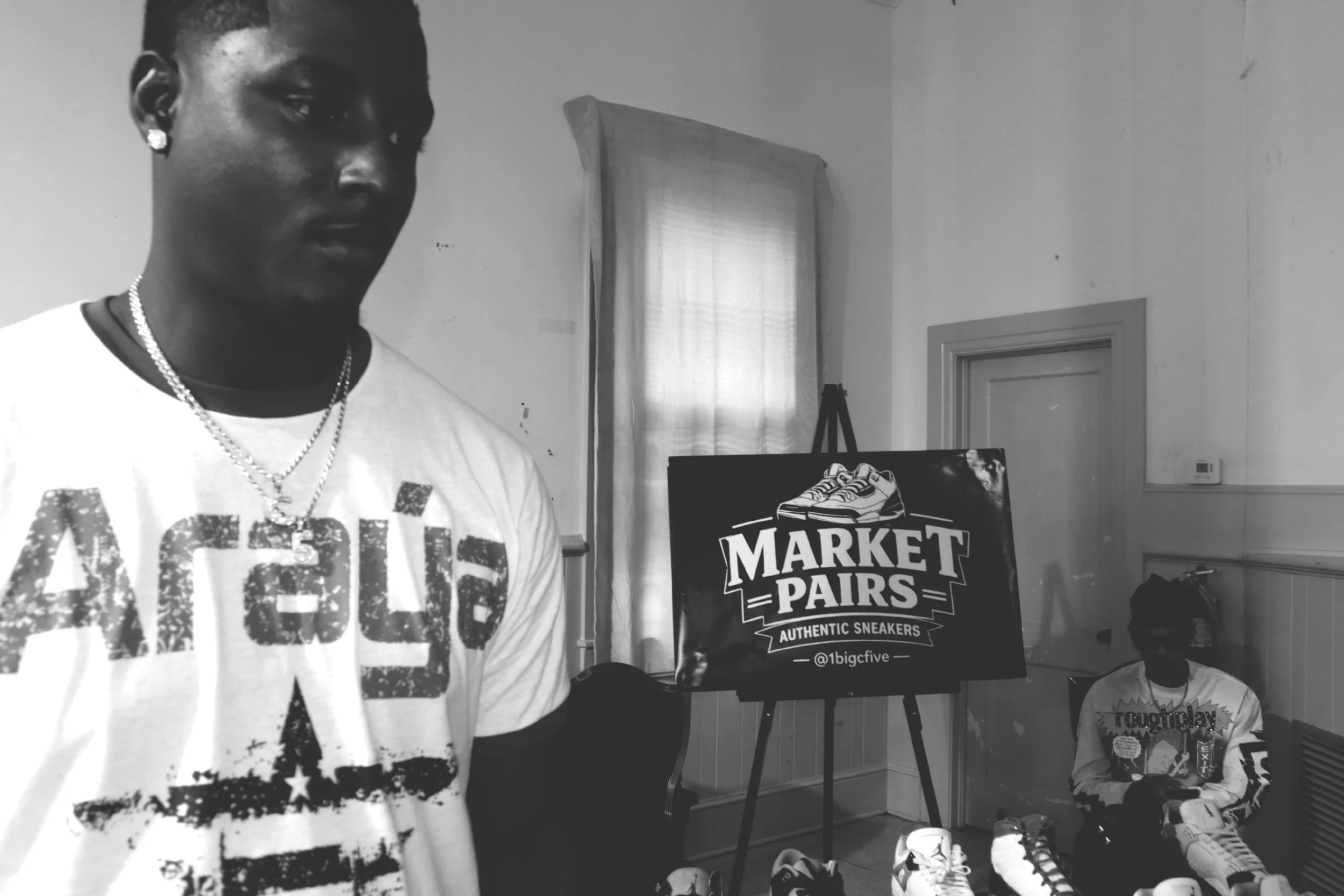 Black and white photo of a person with earrings and necklaces, standing indoors near a table with sneakers and a sign for market pairs.