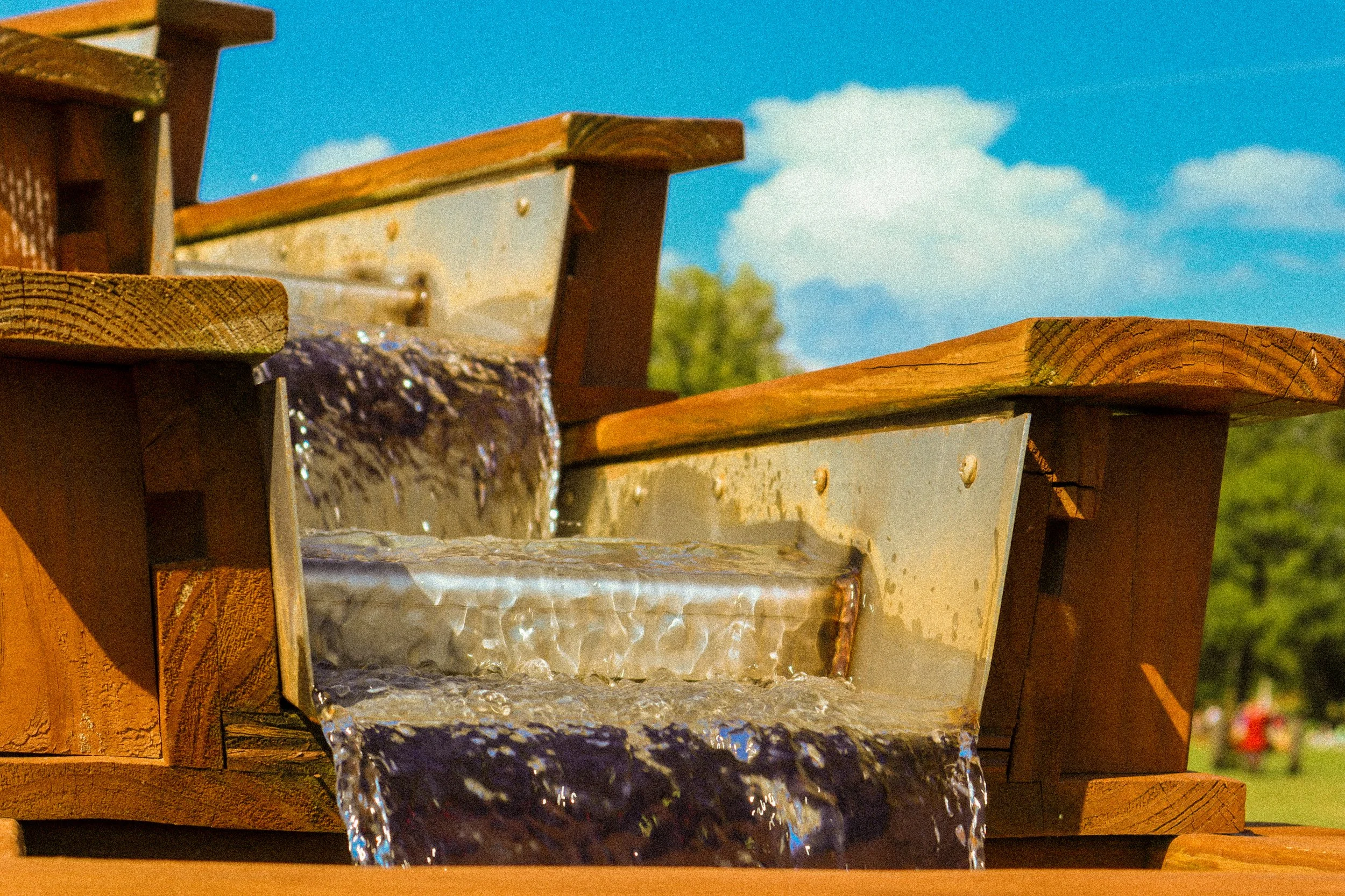 Close-up image of a wooden water fountain with water flowing over textured rocks, set outdoors against a background of blue sky and green trees.