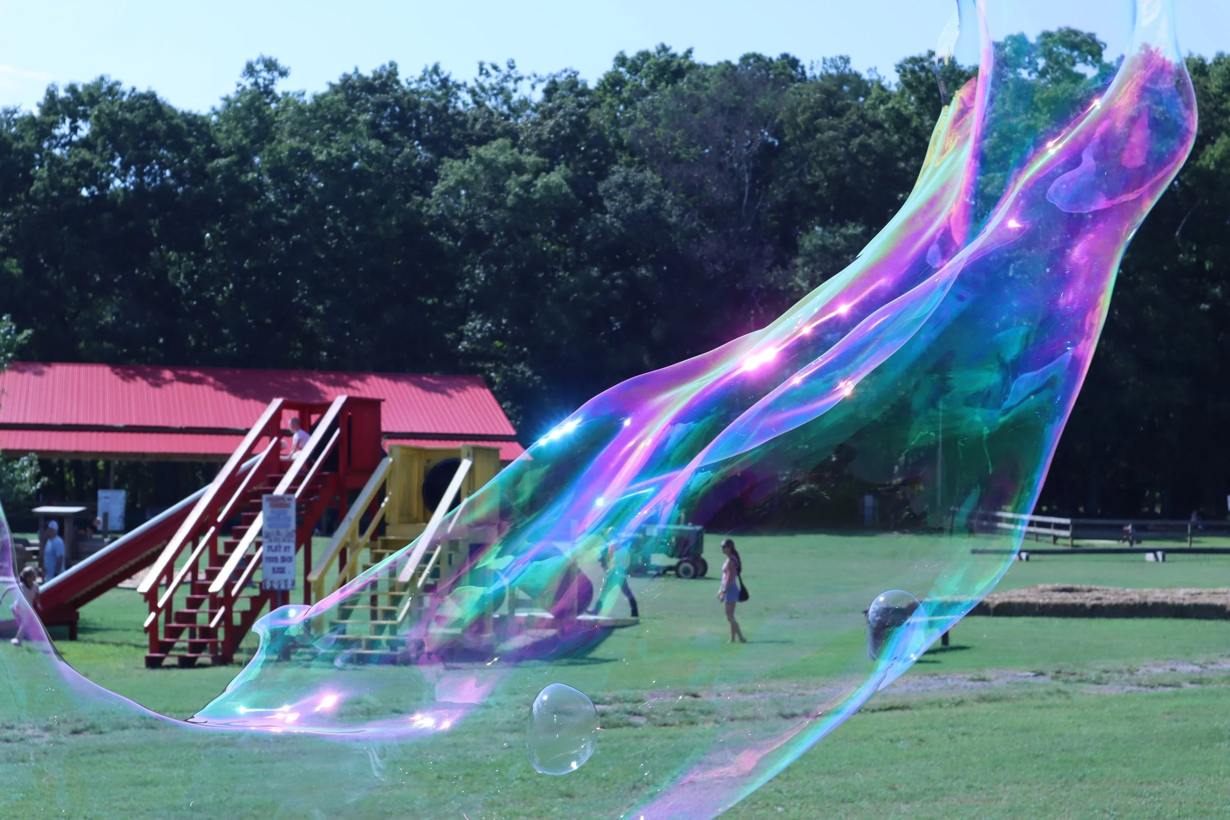 Colorful giant soap bubble floating over a park with playground equipment, children, and green trees in the background.
