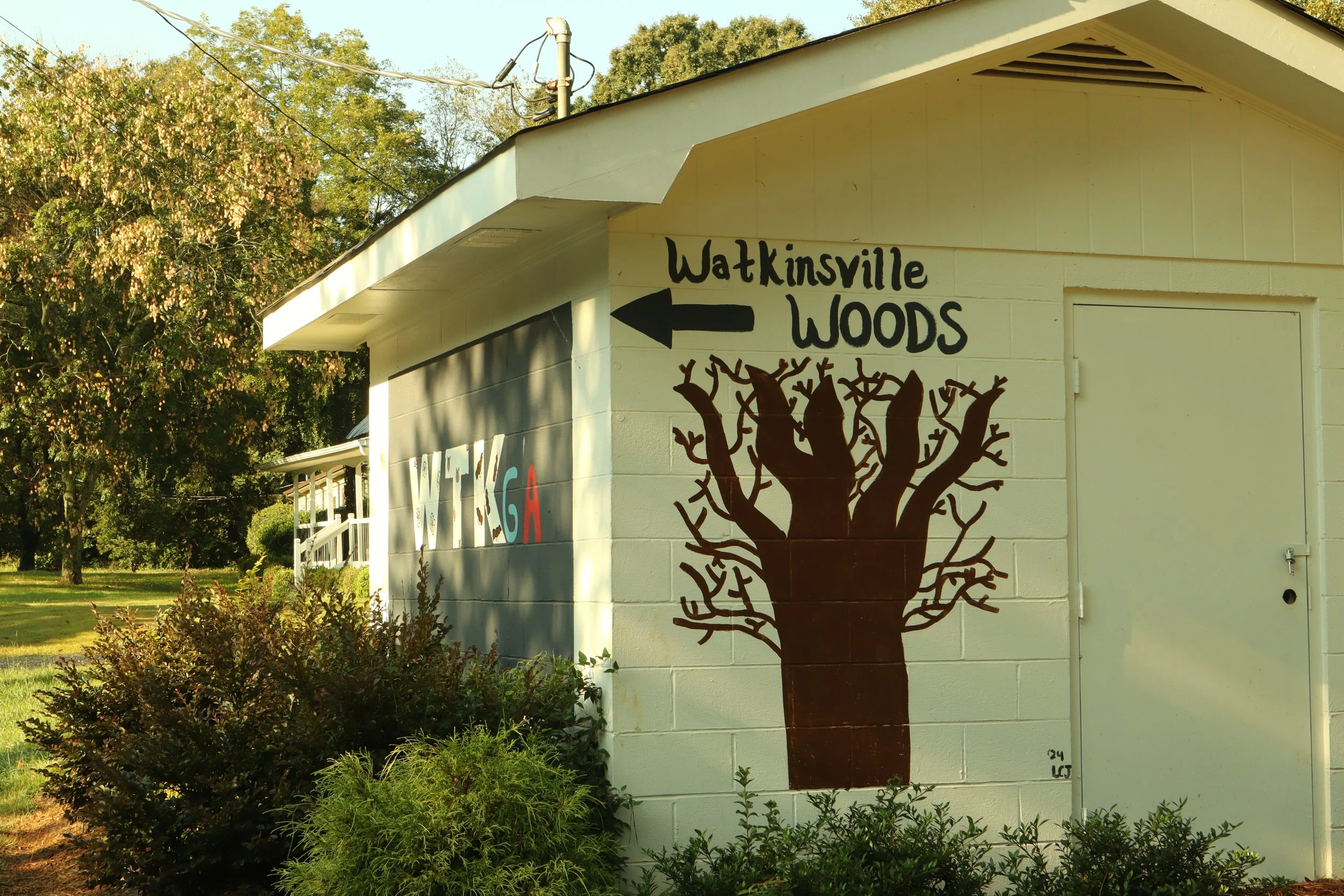 Building with a mural of a tree painted on the front, featuring the text 'Watkinsville Woods' with an arrow pointing left, and surrounding trees and greenery in a sunny outdoor setting.