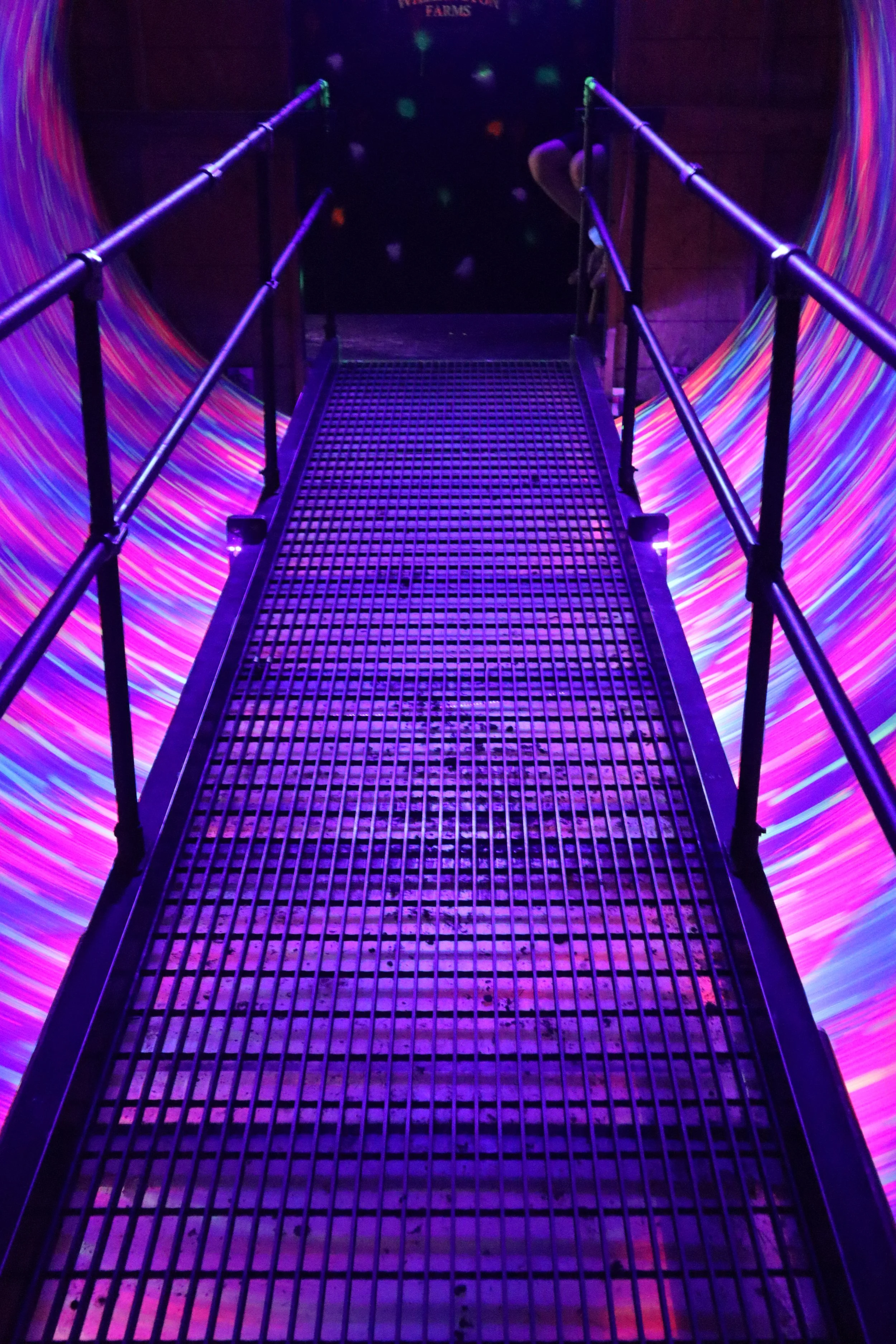 A metal bridge with purple railing in a dark room with colorful, swirling lights on the sides.