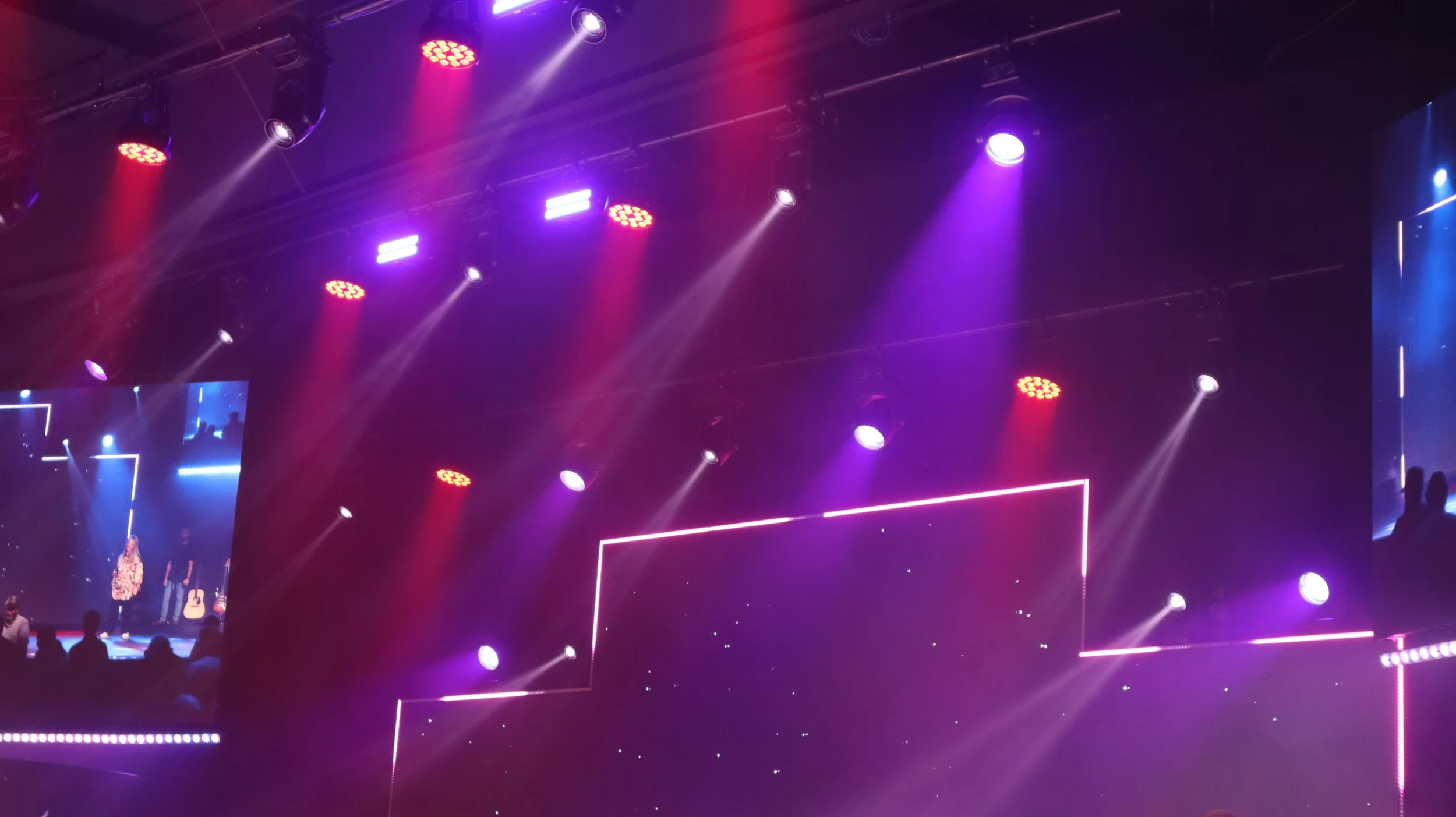 Stage with colorful purple and pink lights, and a large screen showing a performer or speaker, with silhouettes of audience members