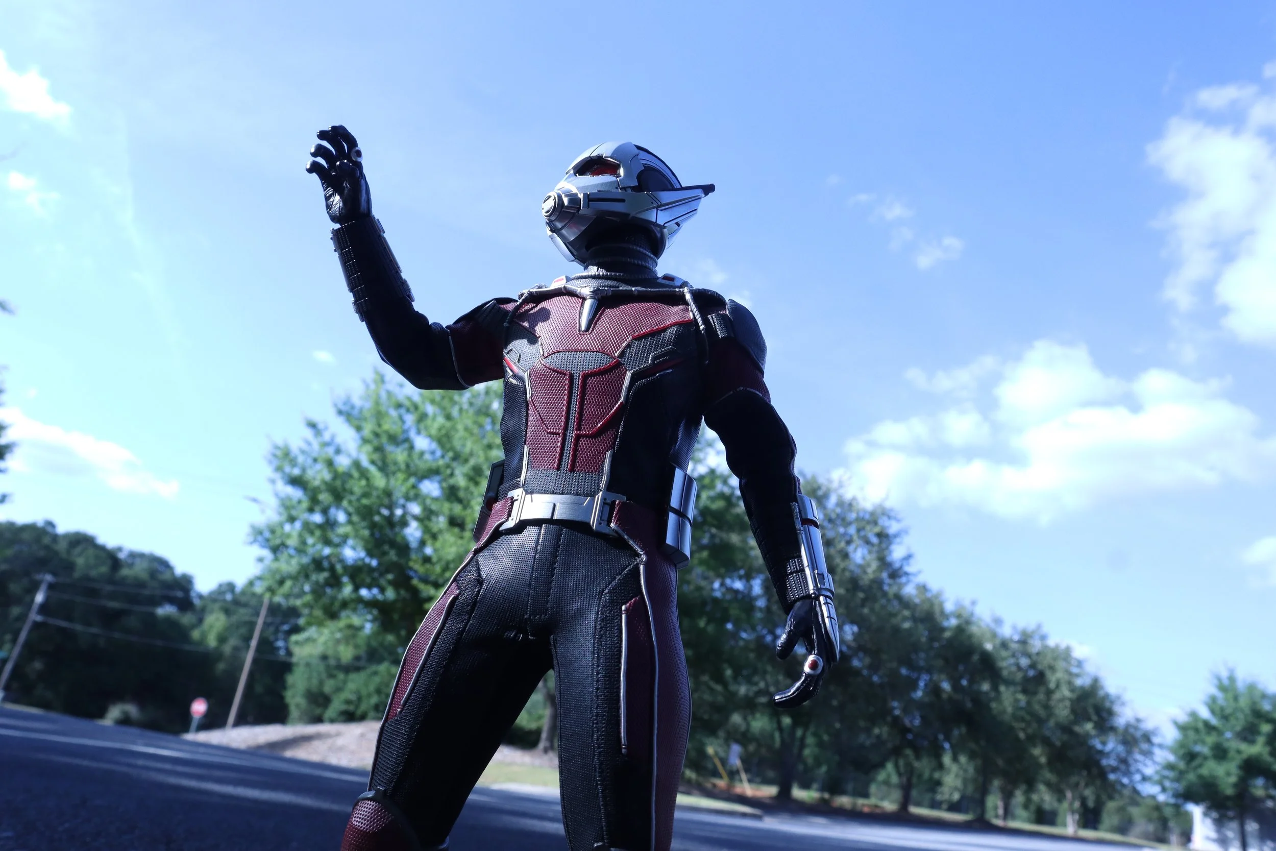 Close-up of a futuristic robot or superhero in a black and red suit, standing outdoors against a clear blue sky with trees in the background, raising one arm.