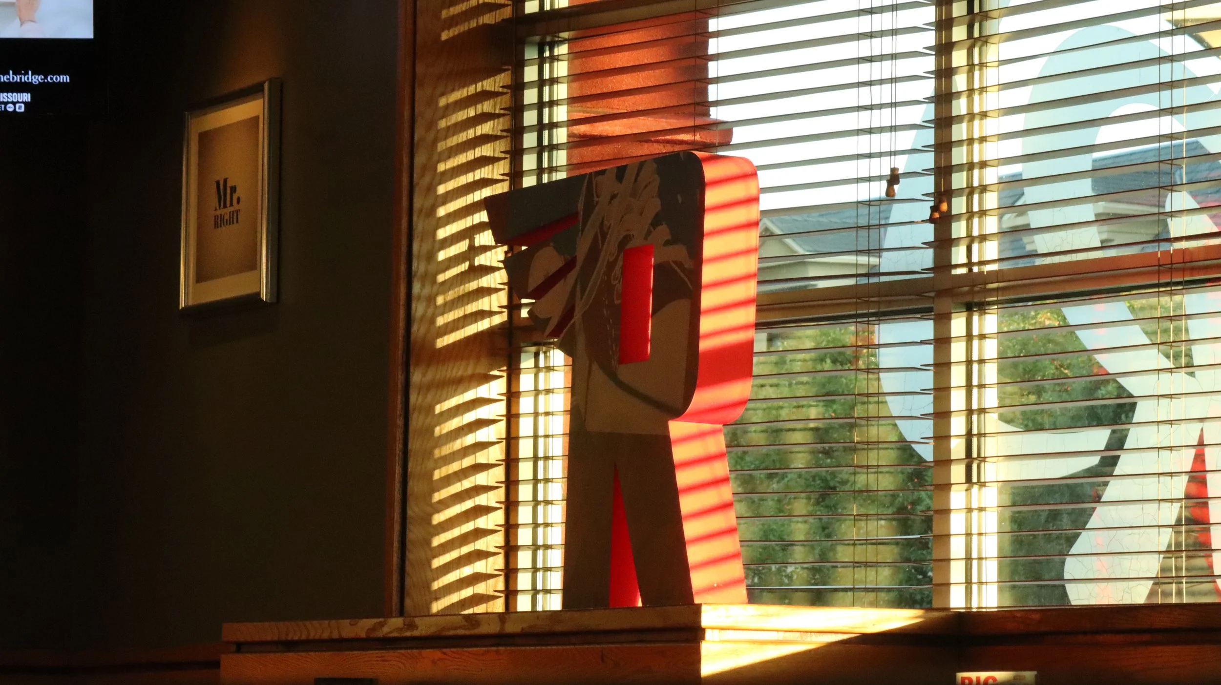 Sunlight streaming through wooden blinds onto a red and white abstract sculpture inside a room with dark walls, a framed sign that reads 'Mr. RIGHT,' and windows overlooking trees and a building outside.