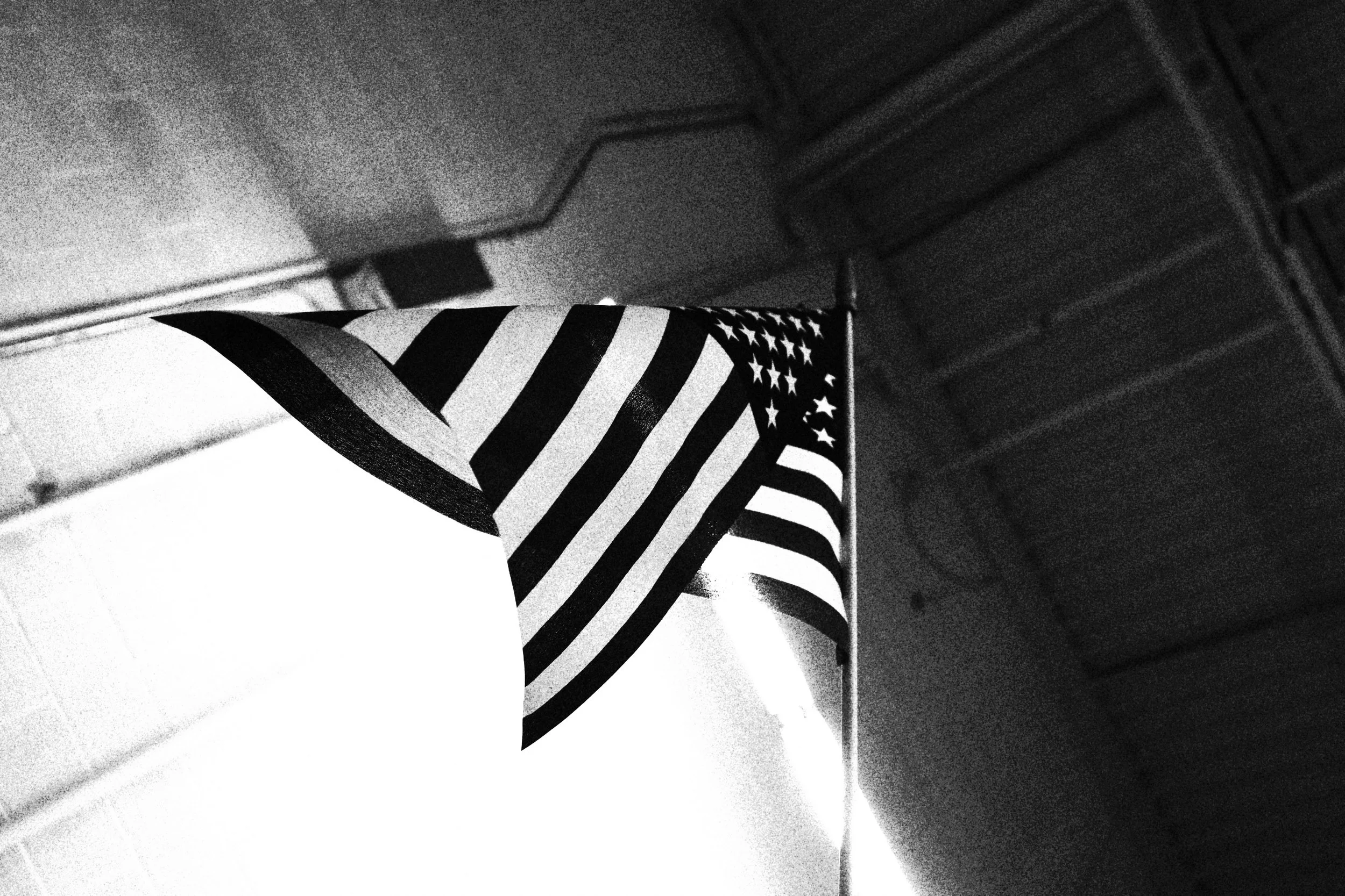 Black and white photograph of an American flag hanging indoors, with a staircase and wall in the background.