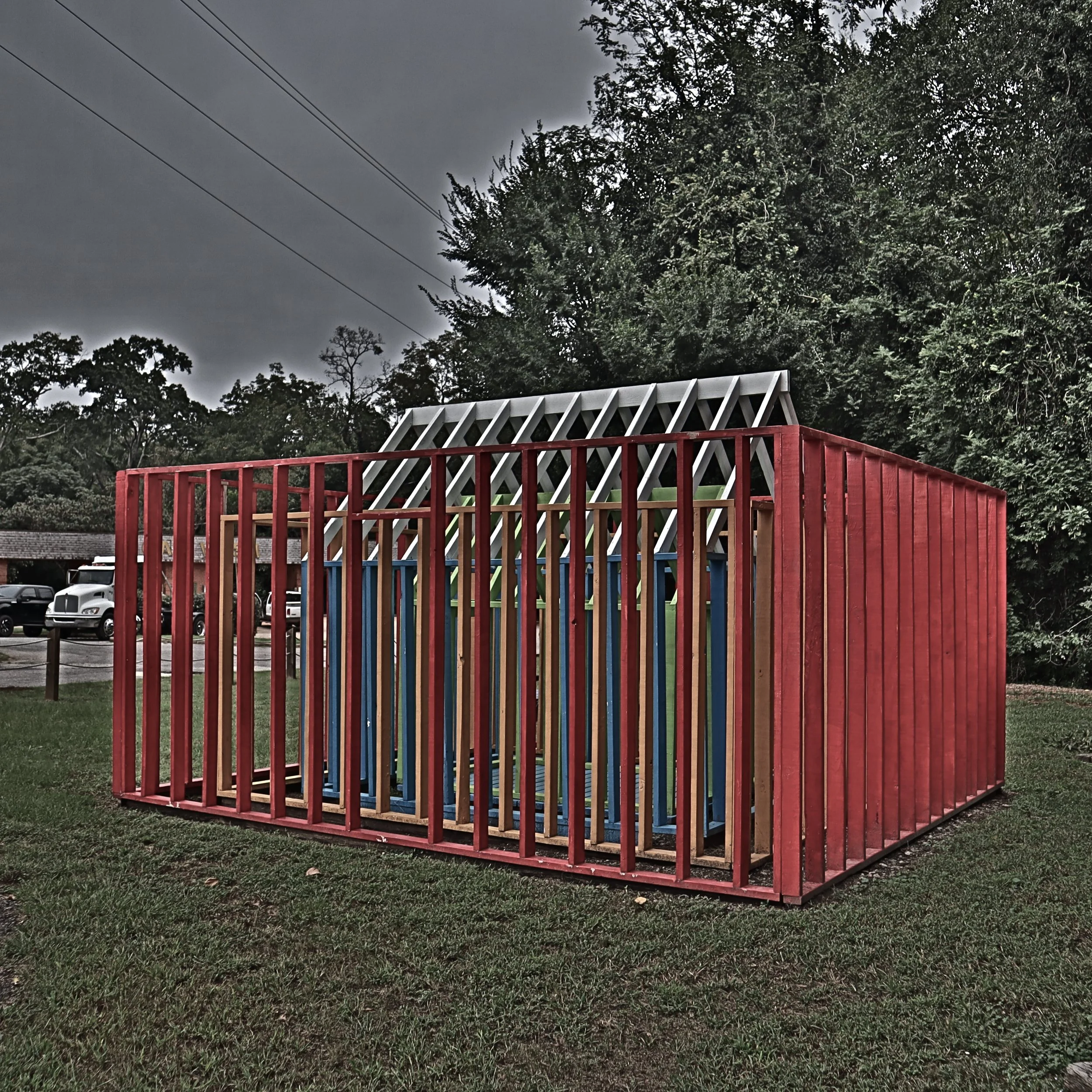 A colorful wooden playground structure with red, blue, green, and gray sections surrounded by grass and trees, with an overcast sky in the background.