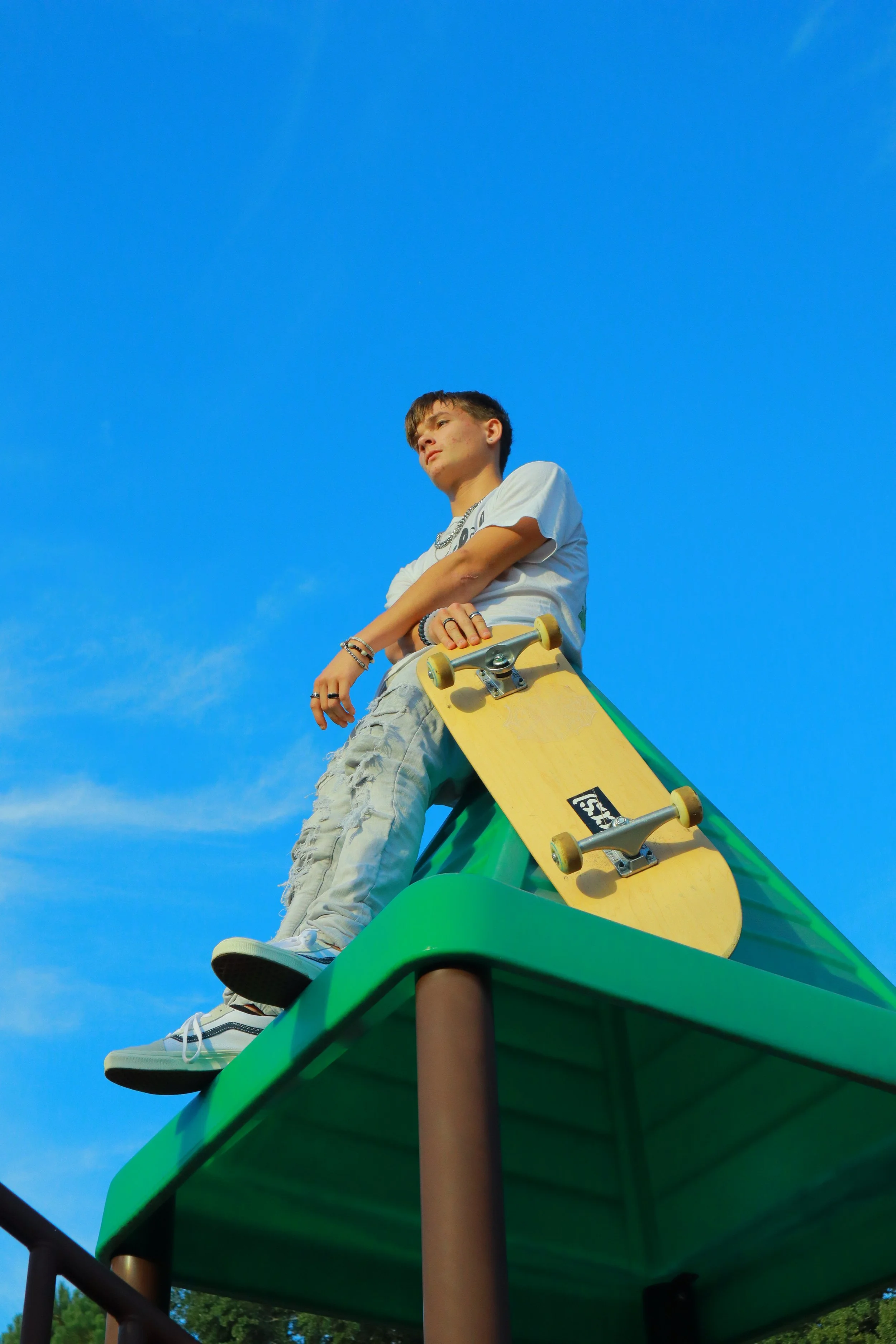 A young person with short brown hair, wearing a white T-shirt and ripped light-colored jeans, sitting on a yellow skateboard at the top of a green playground slide, against a bright blue sky.